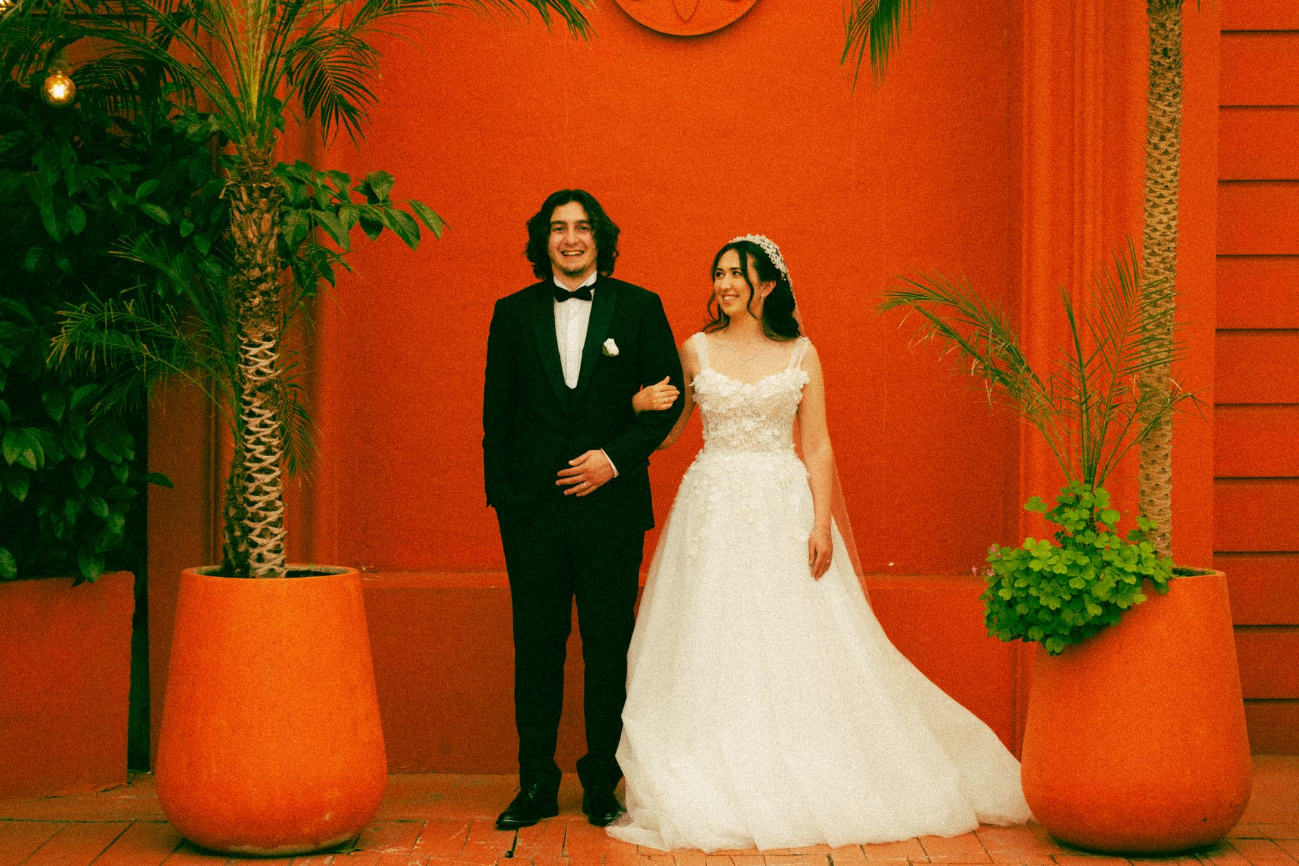 A joyful wedding couple poses against a vibrant orange wall with lush greenery.