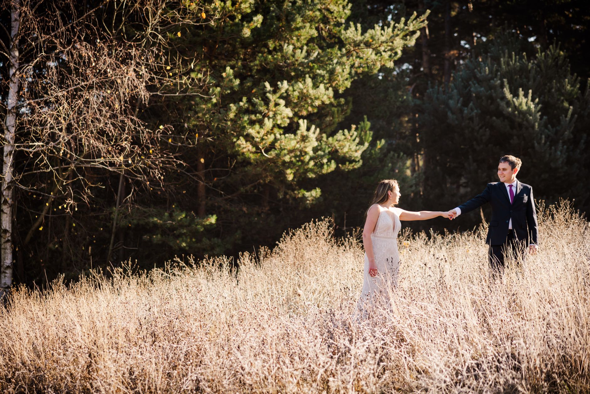 Bride in white gown and groom in suit holding hands in sunlit field.