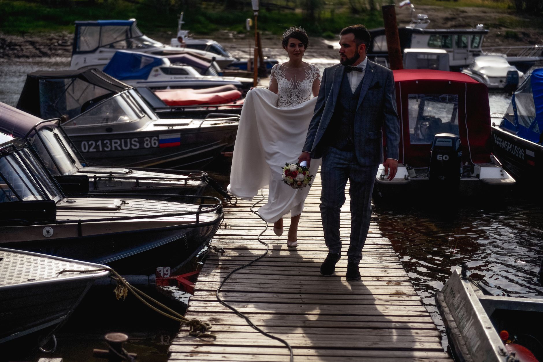 Bride and groom walk hand in hand on a wooden dock surrounded by boats.