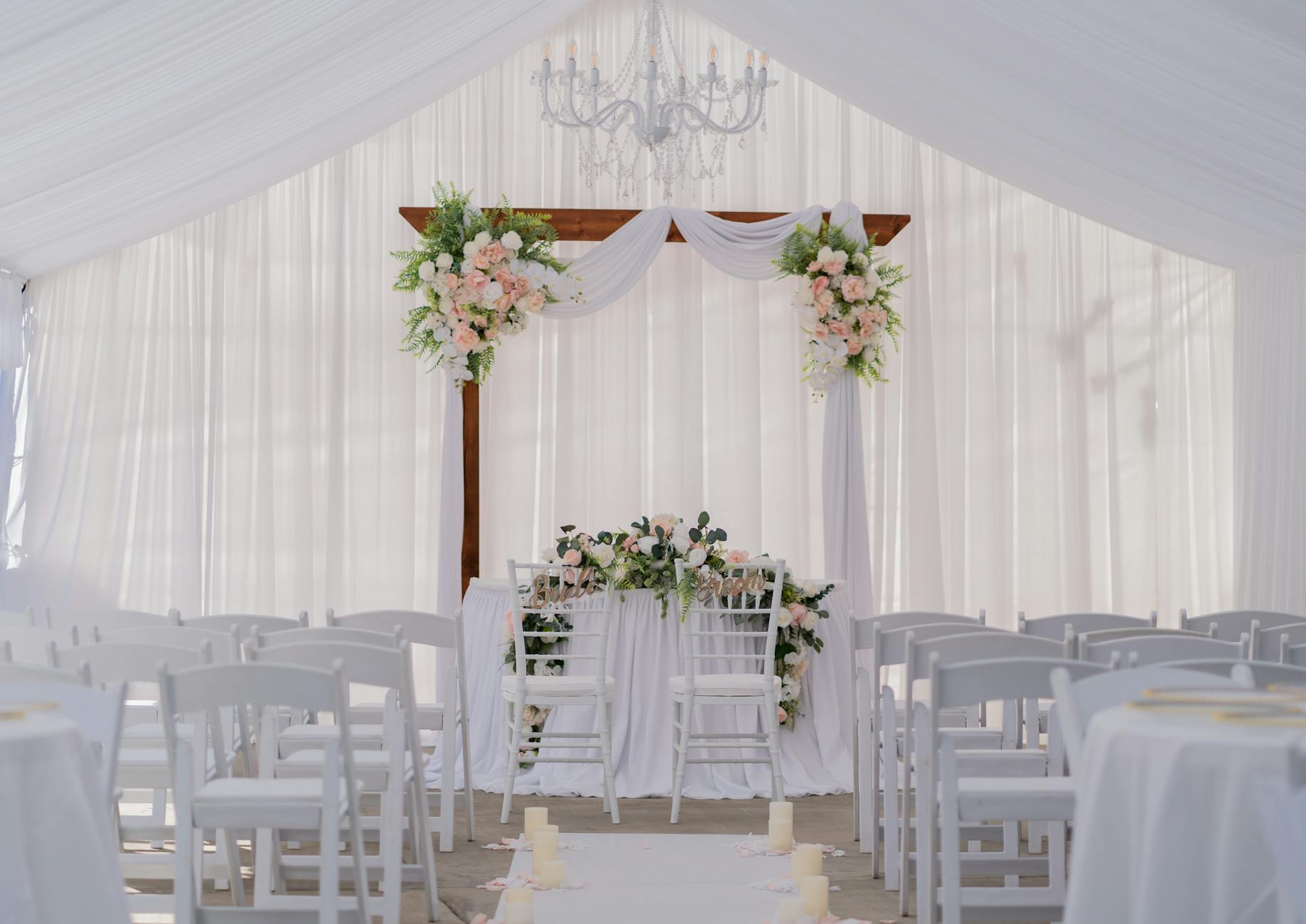 Beautiful wedding ceremony setup with white drapery, floral arrangements, and chandelier. Perfect for elegant nuptials.