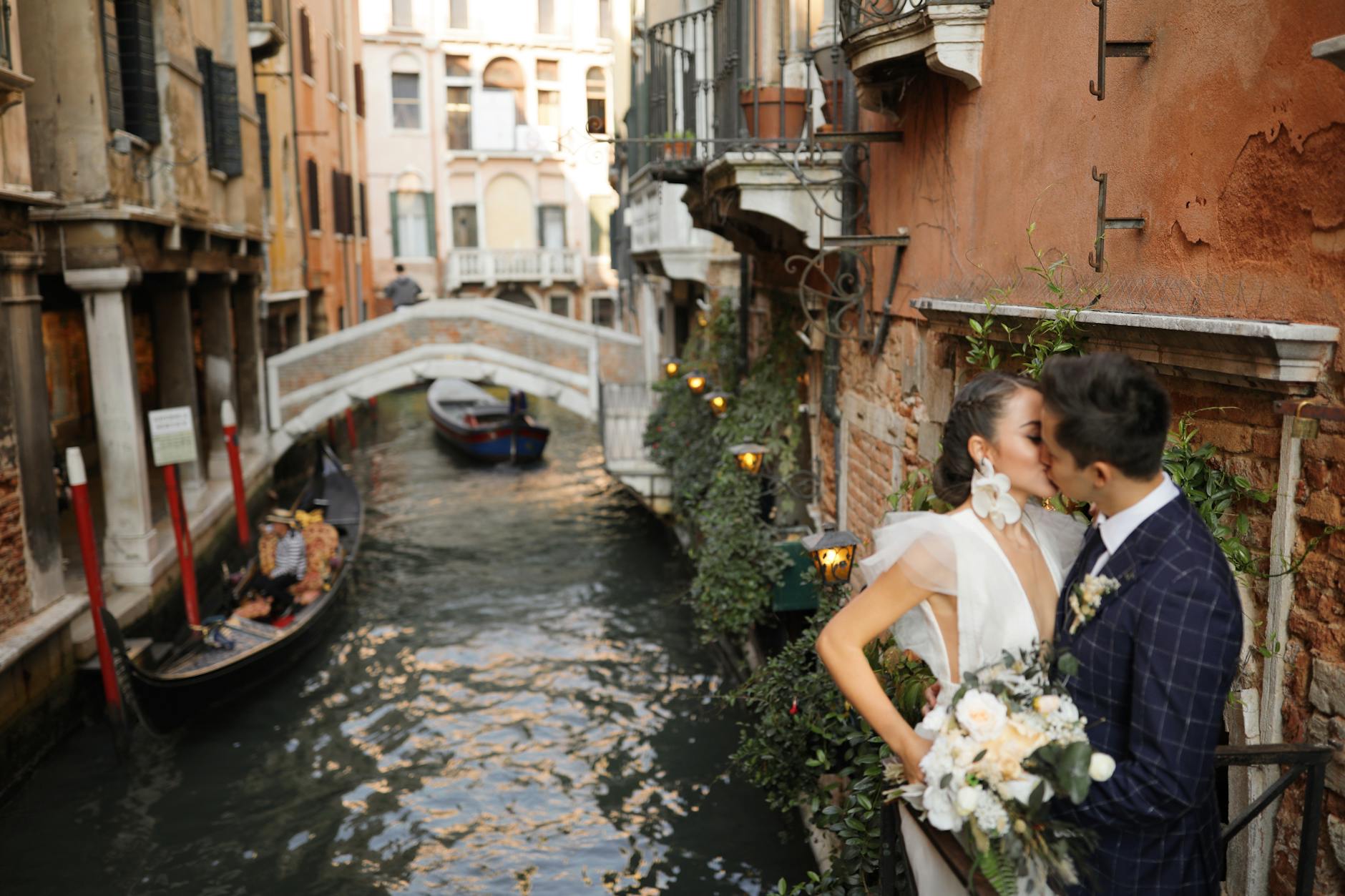 Bride and groom share a kiss beside a picturesque canal in Venice, Italy, with a gondola in the background.