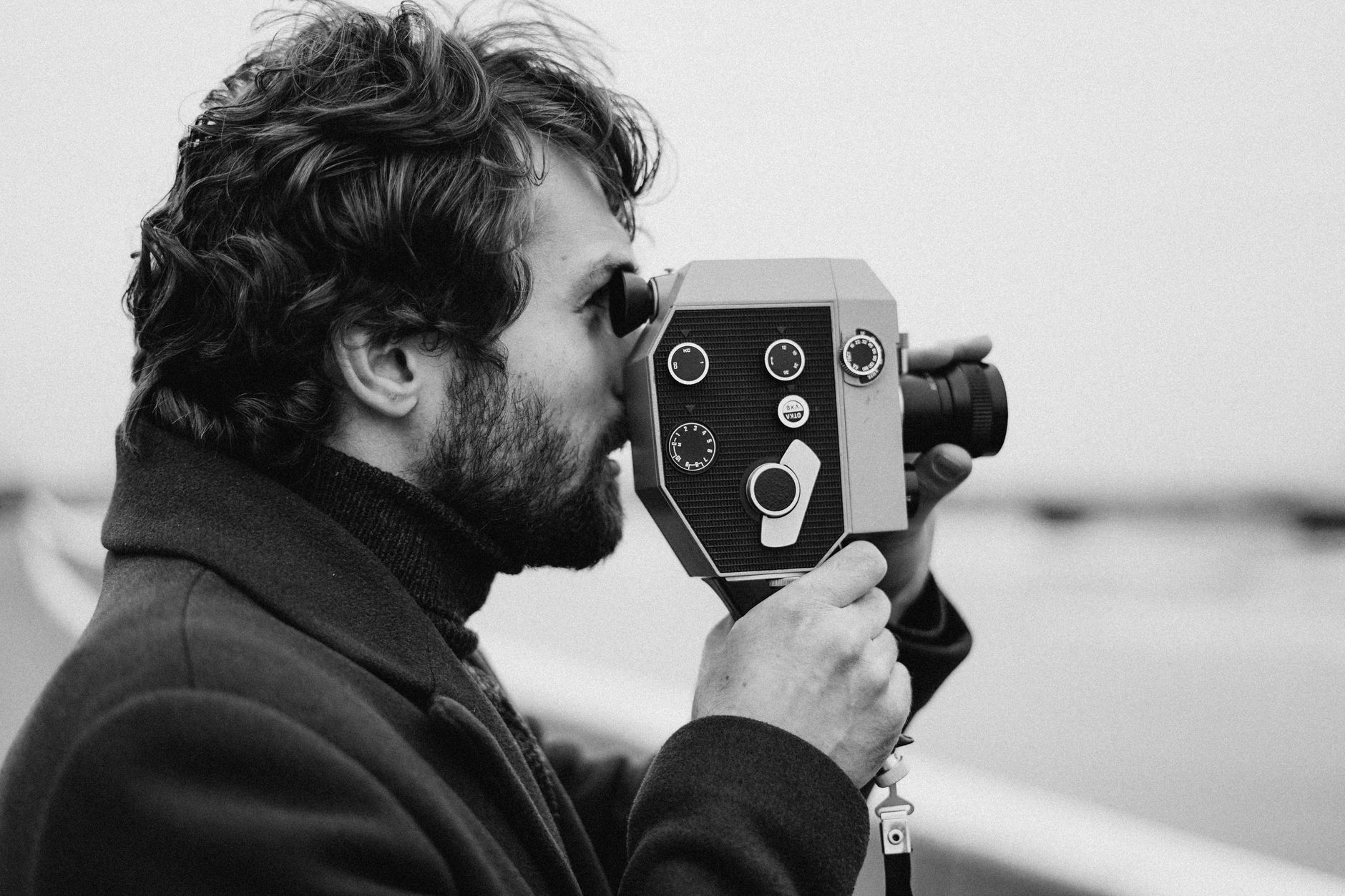 Side view of a man filming with a vintage camera in a black and white setting.