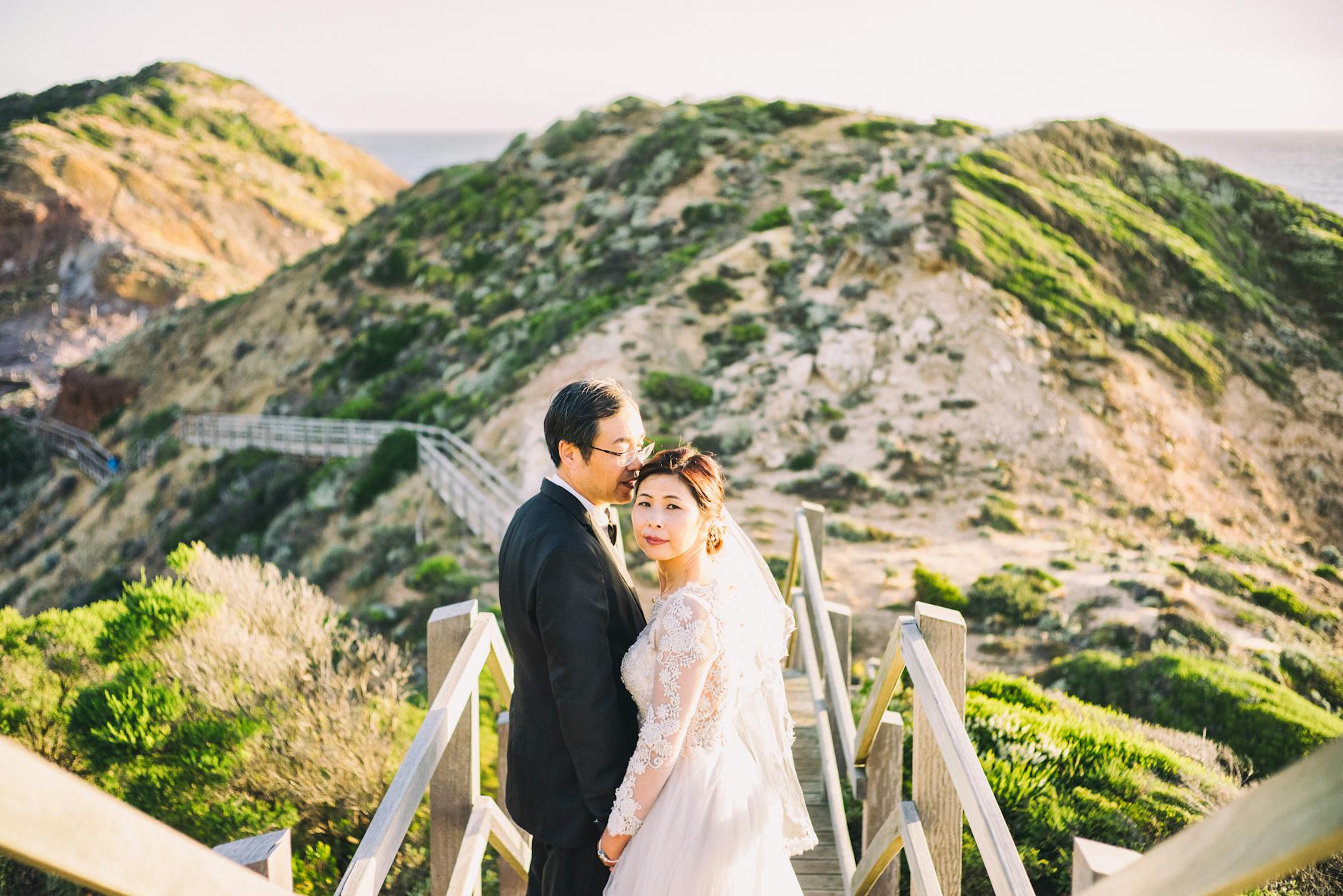 A bride and groom pose on a scenic walkway with expansive mountain and ocean views.
