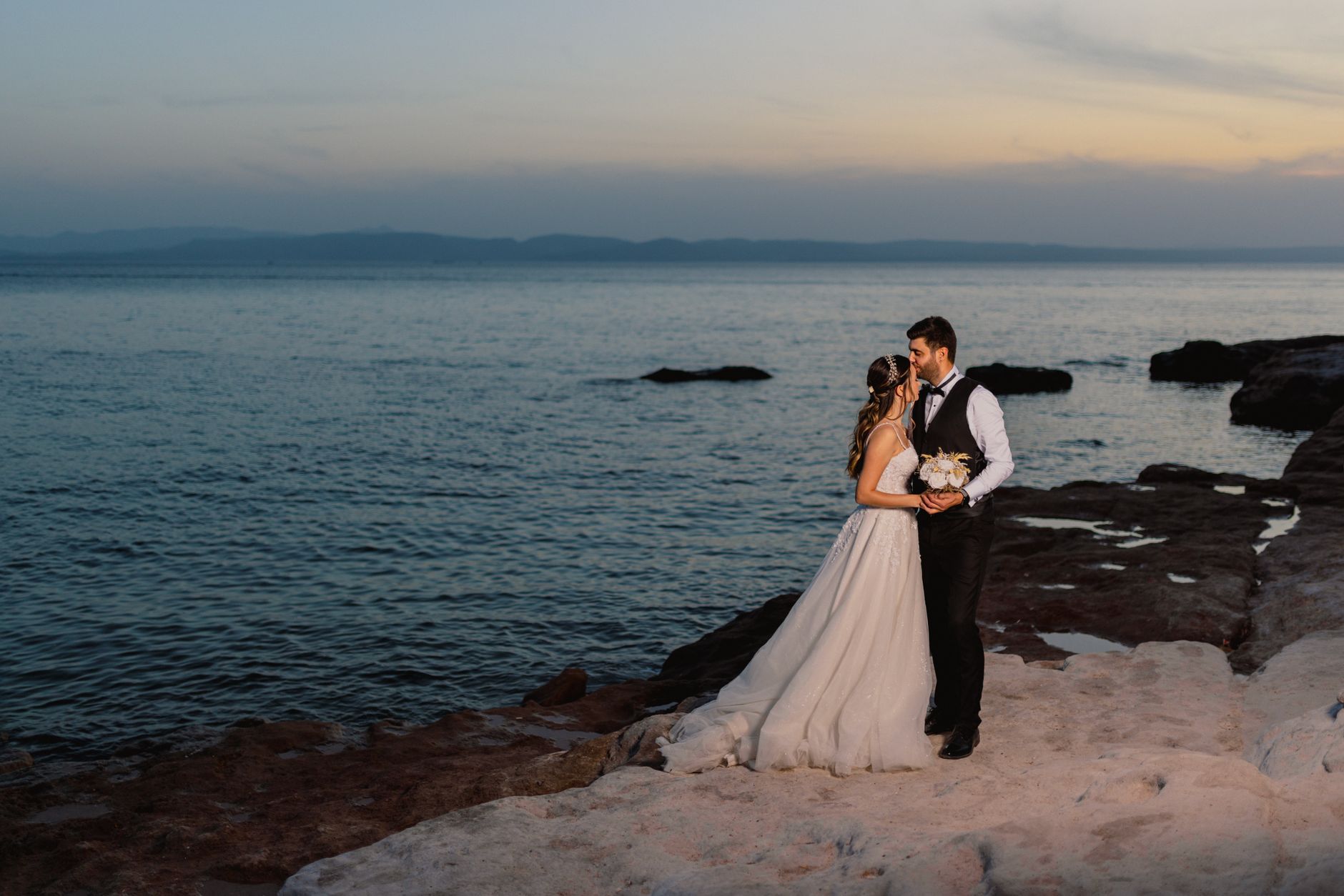 Bride and groom embrace on rocky shore at sunset, capturing love and scenic beauty.