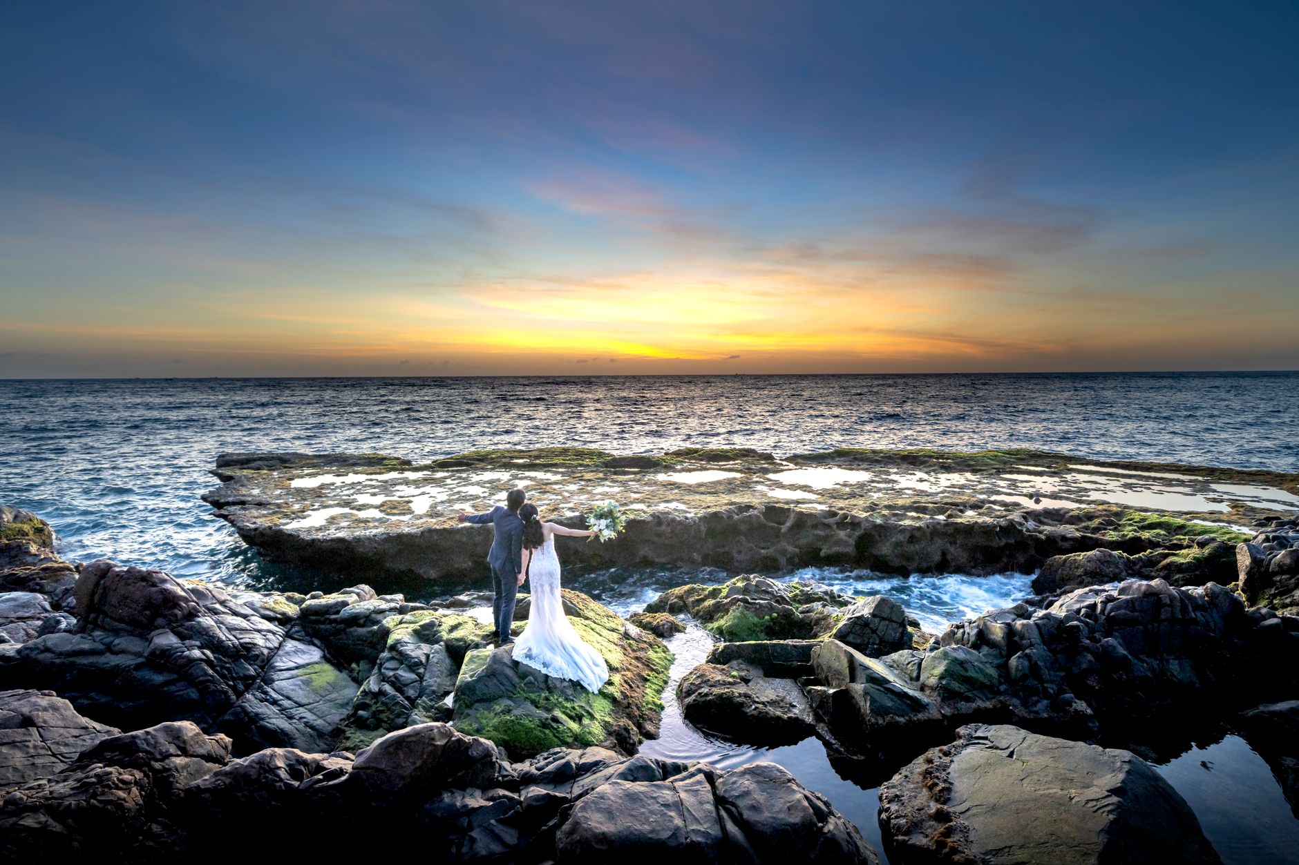 Couple embracing on rocky coast during a stunning sunset backdrop, perfect for wedding themes.
