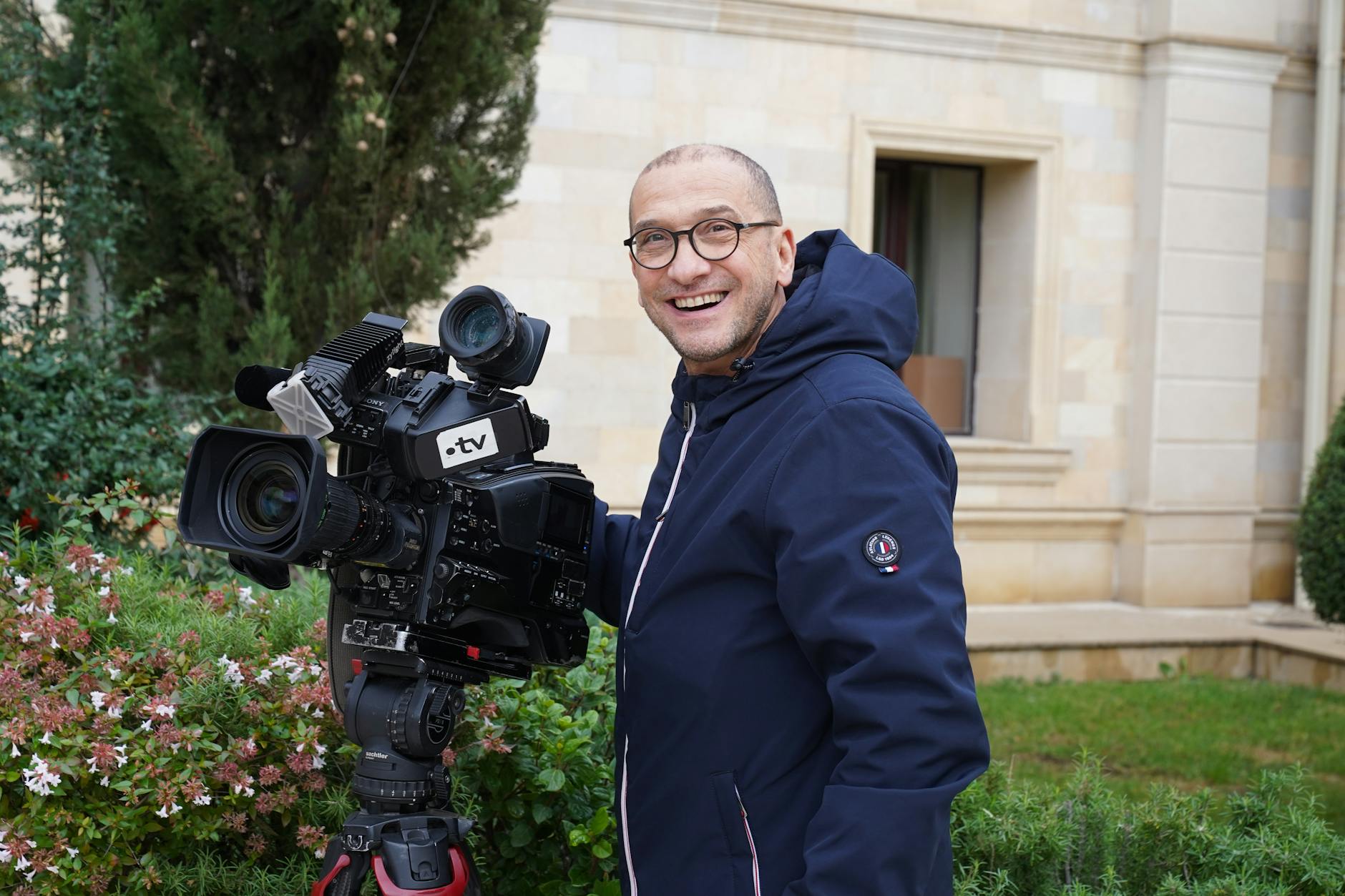 Smiling cameraman with a large TV camera in an outdoor setting near a building.