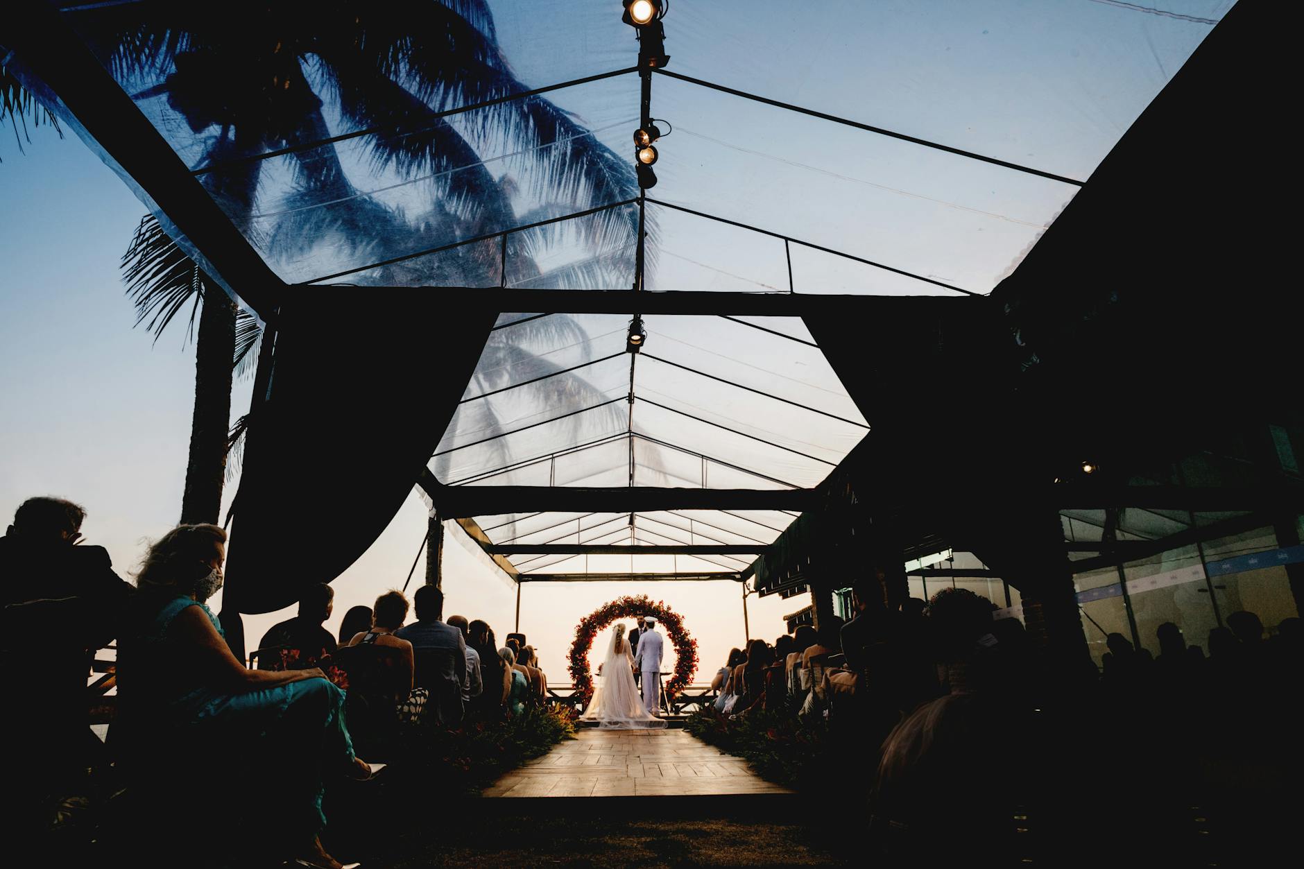 Beautiful evening wedding ceremony under a glass tent with greenery and guests seated.