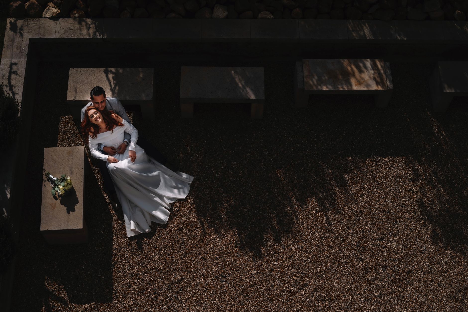 Aerial view of a newlywed couple embracing on a stone bench in a secluded outdoor area.