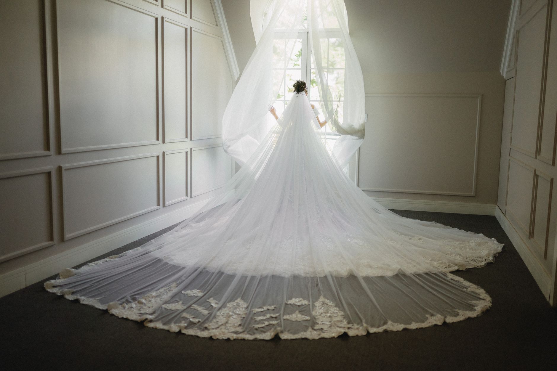 A stunning bride in an elegant gown stands by a sunlit window, creating a serene and ethereal scene.