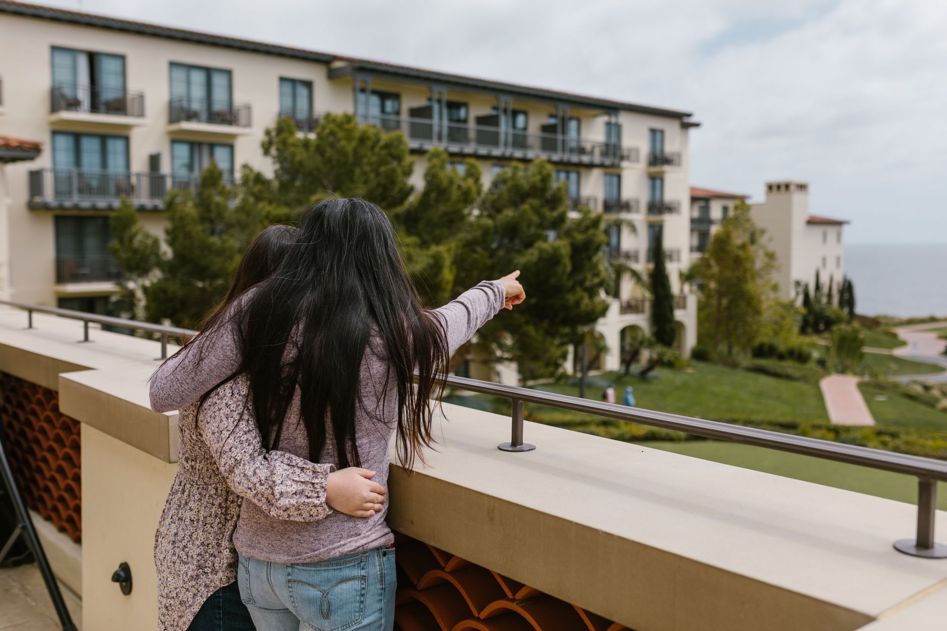 Mother and daughter share a close moment on a balcony, pointing at a scenic view.