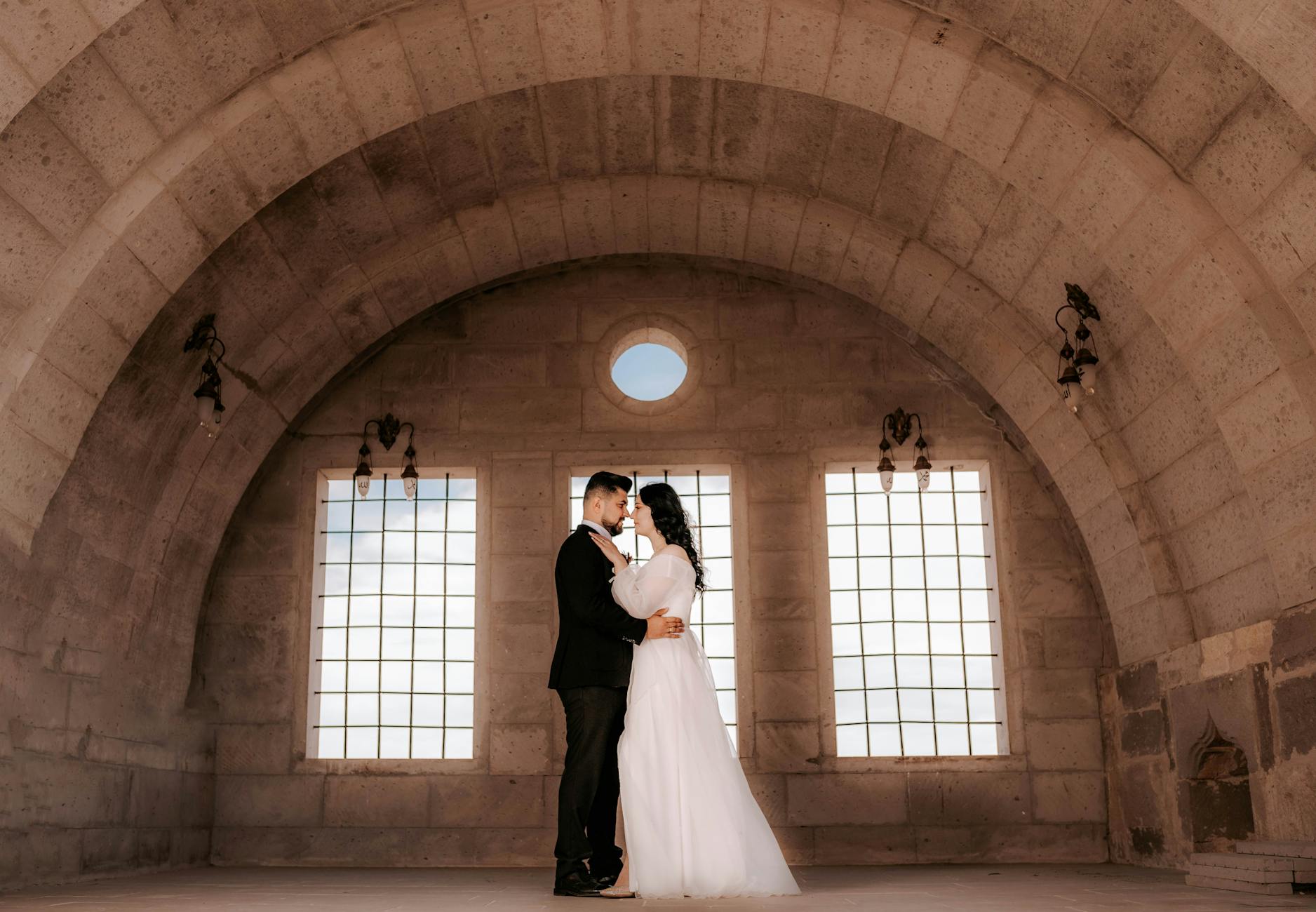 A couple elegantly dressed in wedding attire embracing in a historic stone archway room.