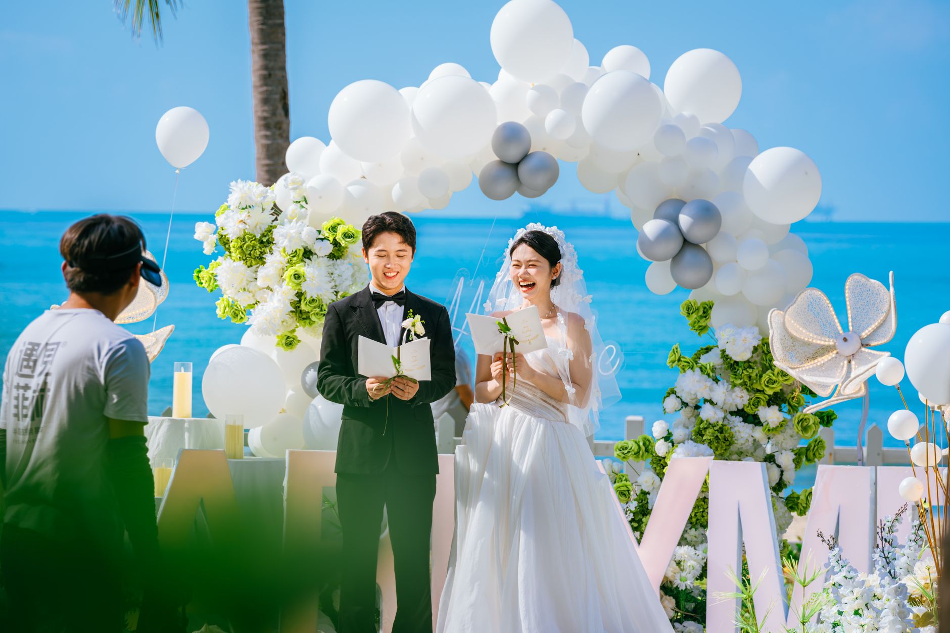 Bride and groom joyfully reciting vows at a scenic beach wedding ceremony under a balloon arch.