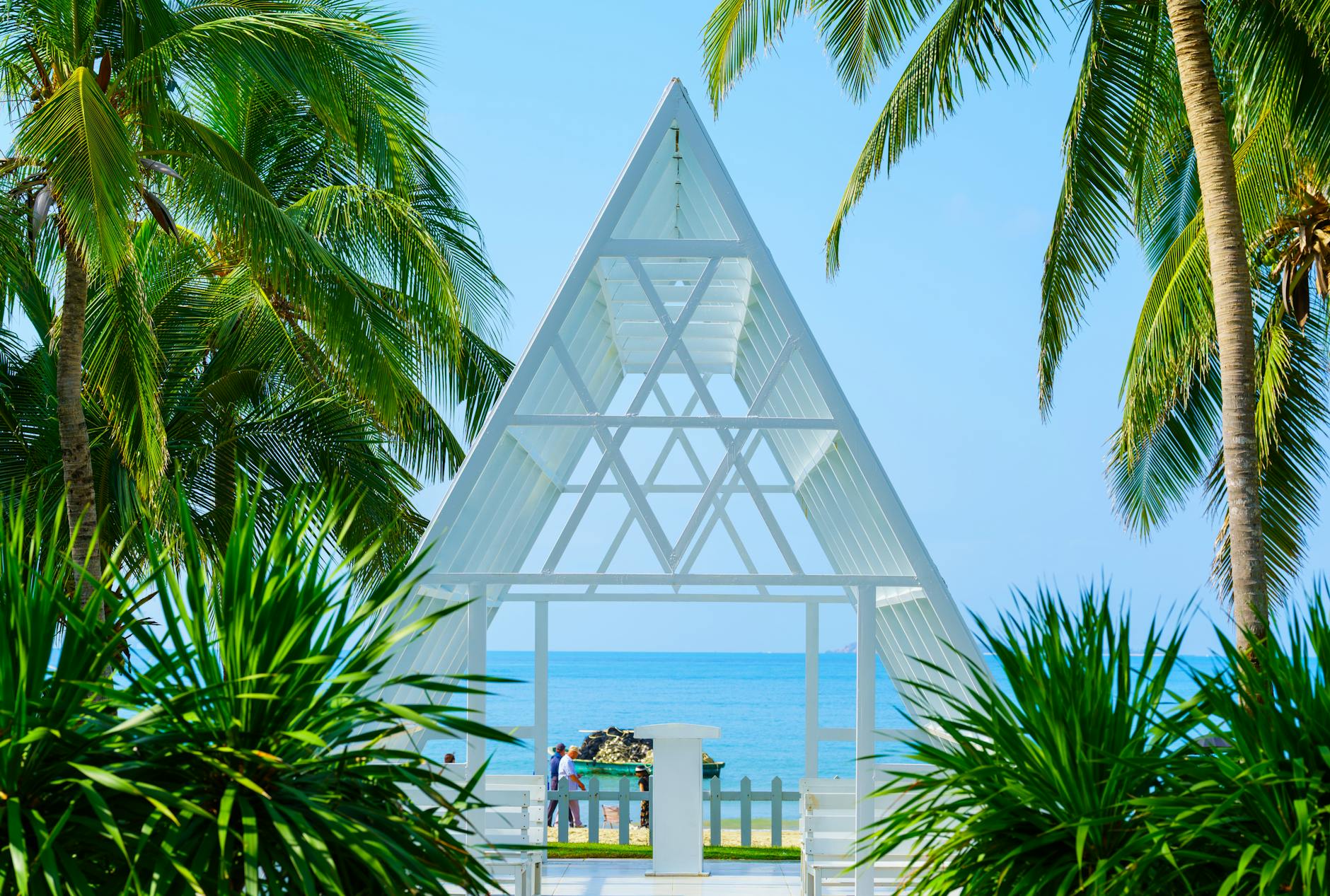 A serene tropical beachfront with a modern A-frame chapel surrounded by palm trees, perfect for weddings.