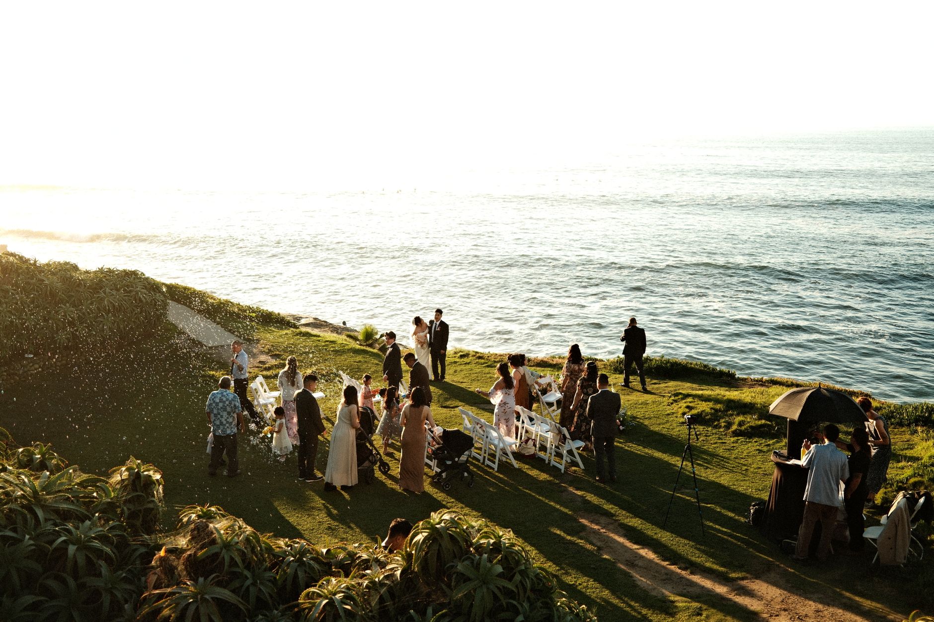 Elegant seaside wedding ceremony with guests gathered on a cliffside during sunset.