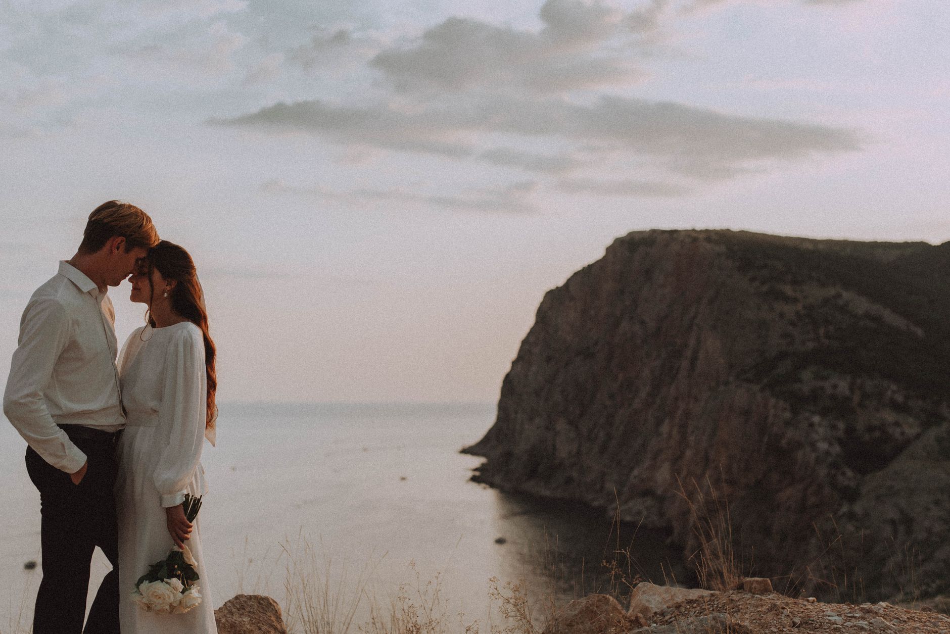 A loving couple embraces on a stunning cliffside during a serene sunset.