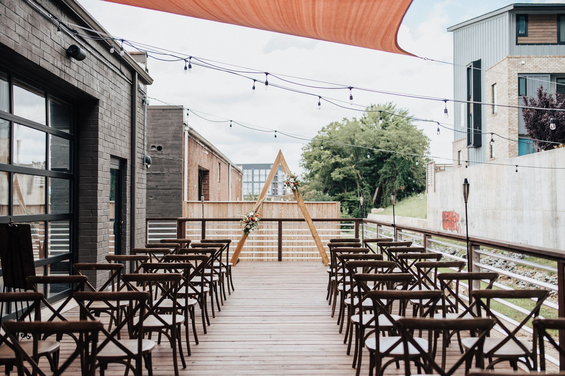 Elegant terrace wedding setup with wooden chairs and triangular arch framing a serene outdoor backdrop.