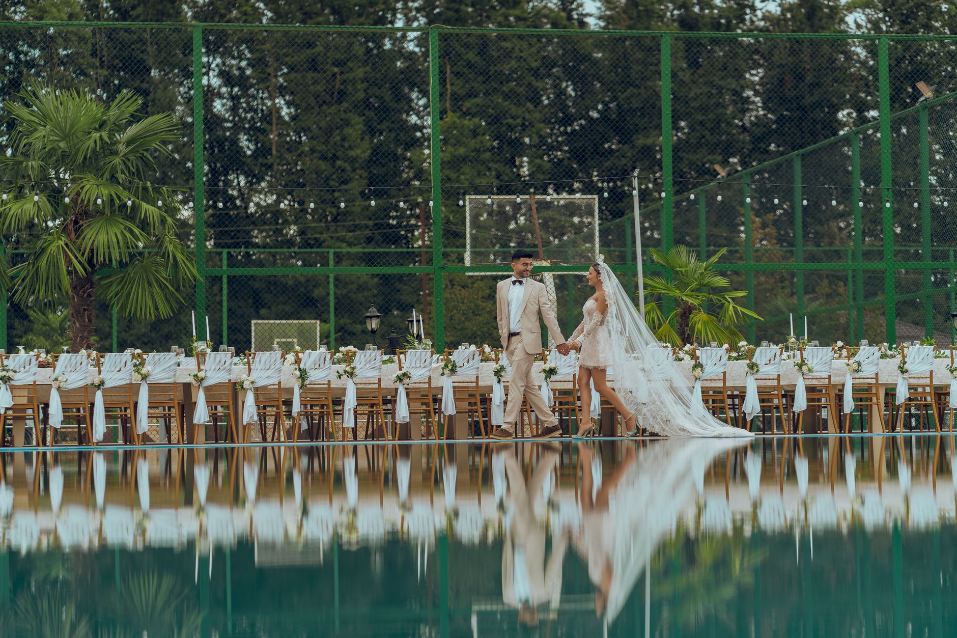 Stylish bride and groom at an outdoor wedding reception by a reflective pool.
