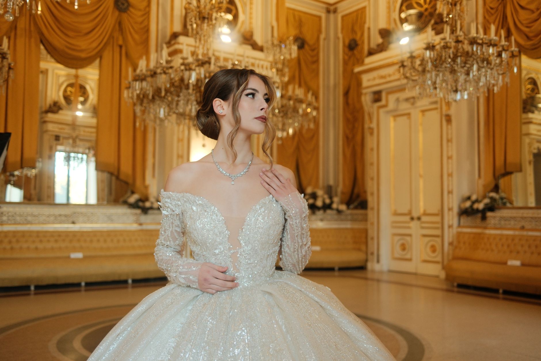 A stunning bride in a glamorous ballroom with chandeliers and golden drapery.