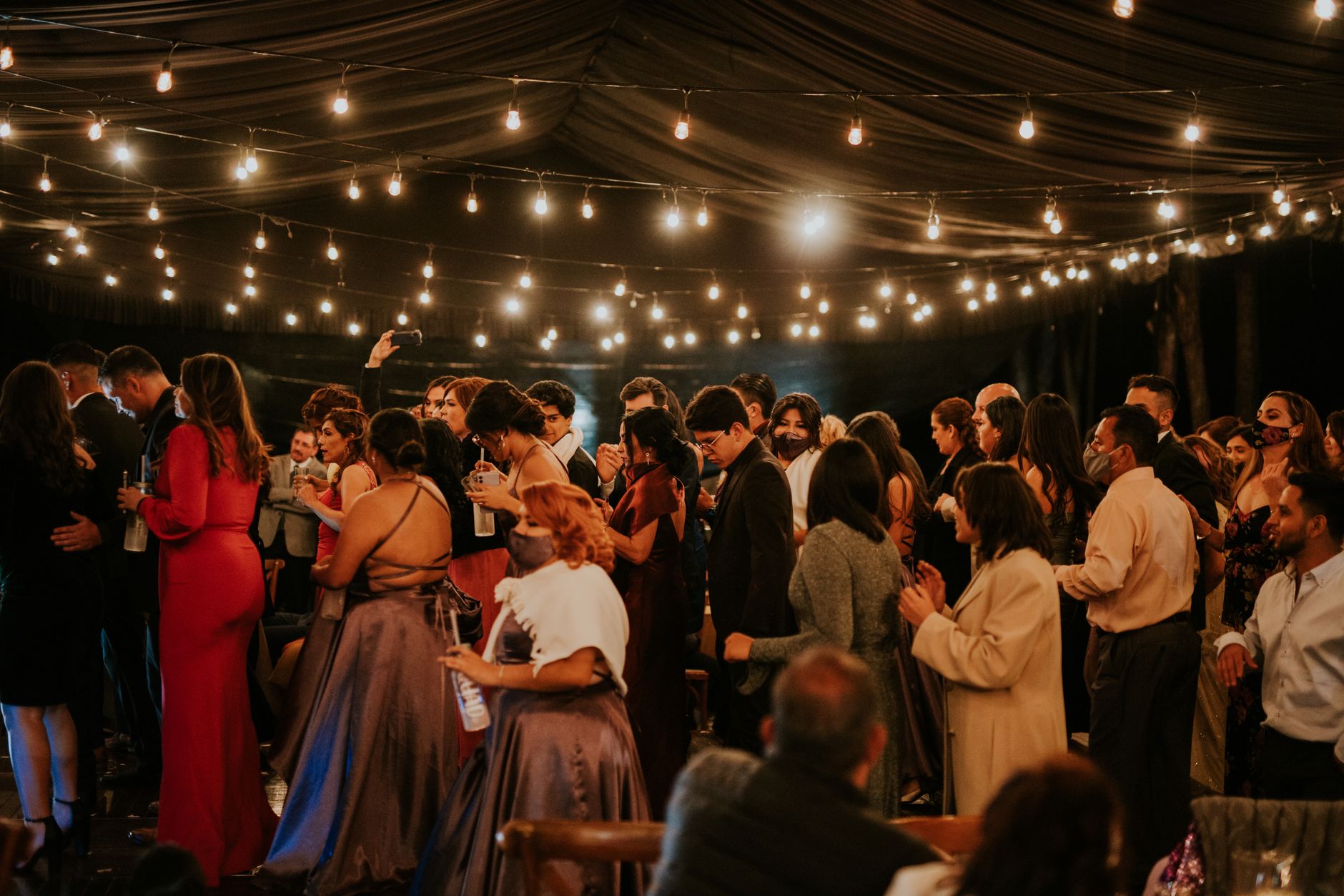 A joyful indoor wedding celebration with elegantly dressed guests under twinkling string lights.