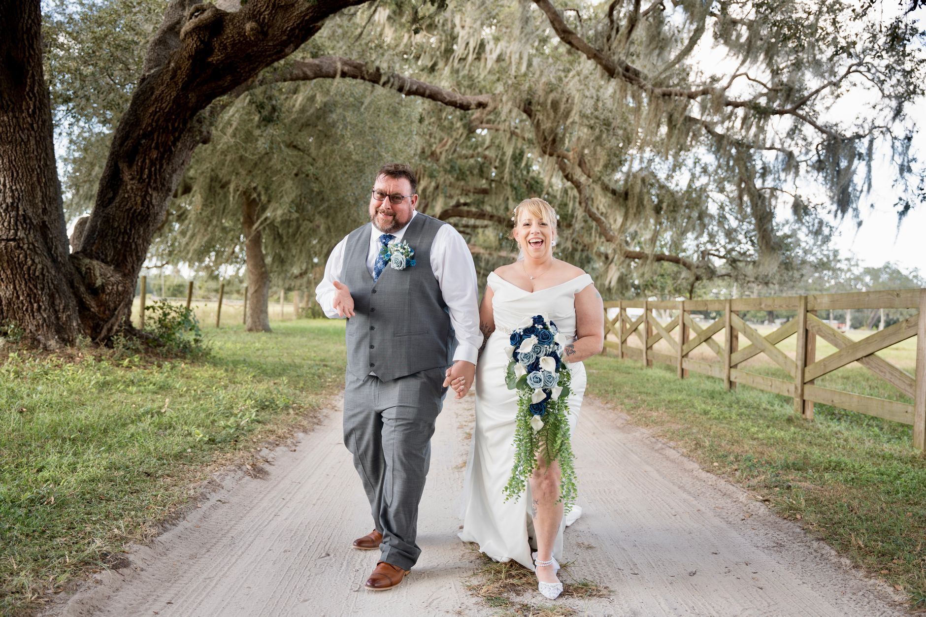 Happy couple walks under lush trees, holding hands on their wedding day.