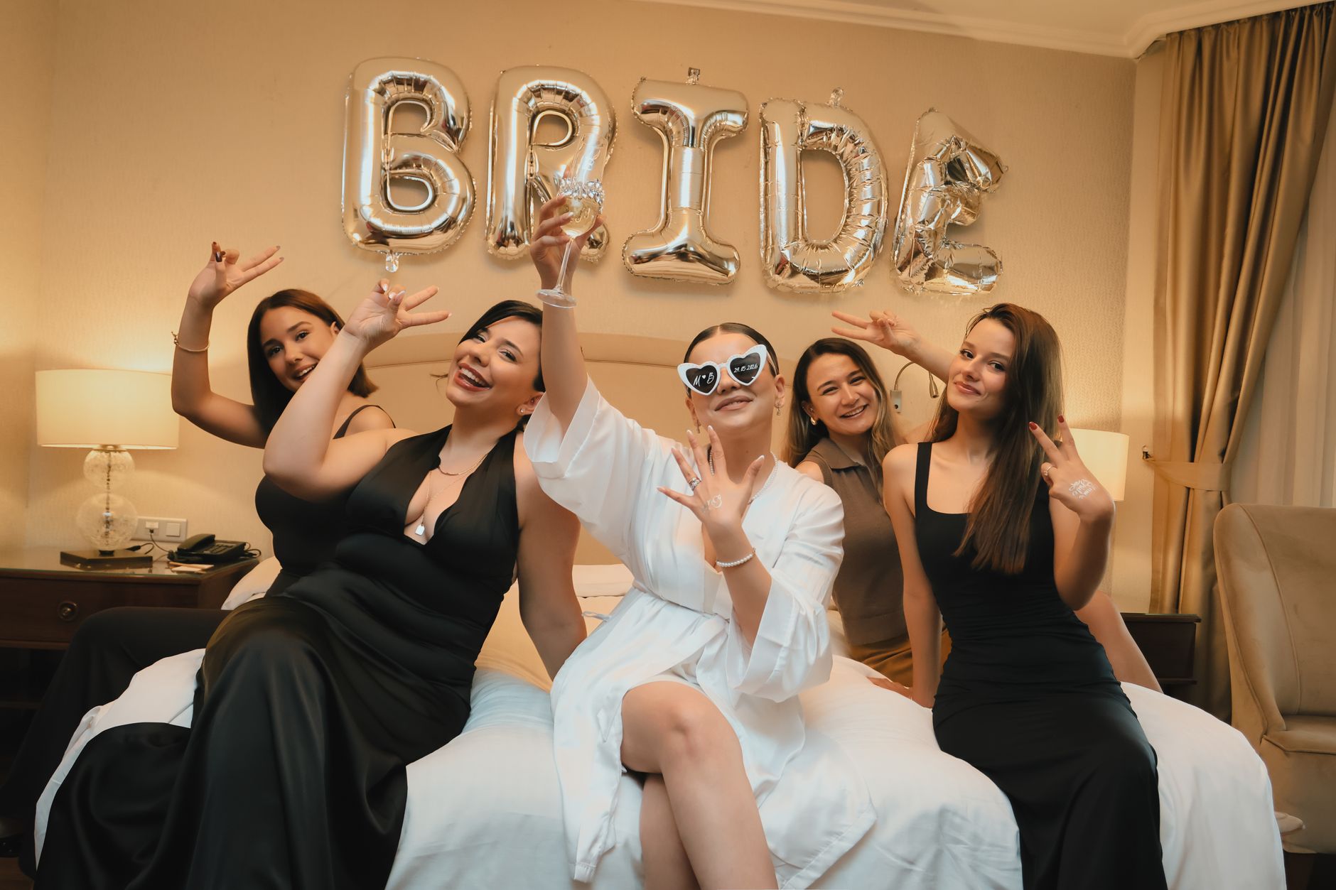Bride and bridesmaids celebrating in a hotel room with balloons spelling 'BRIDE'.