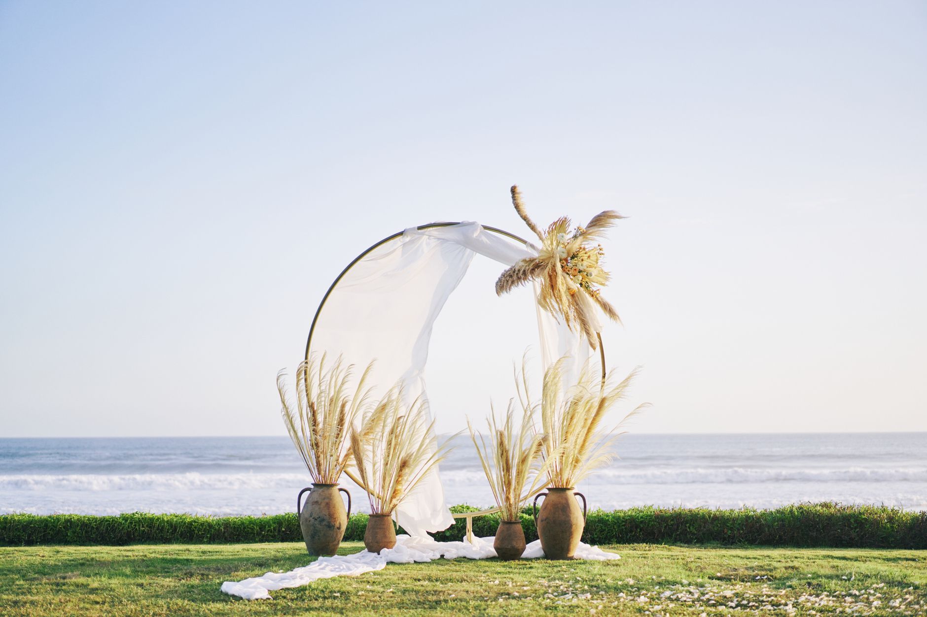 A beautifully decorated beachside wedding arch with pampas grass and vases, set against the ocean backdrop.