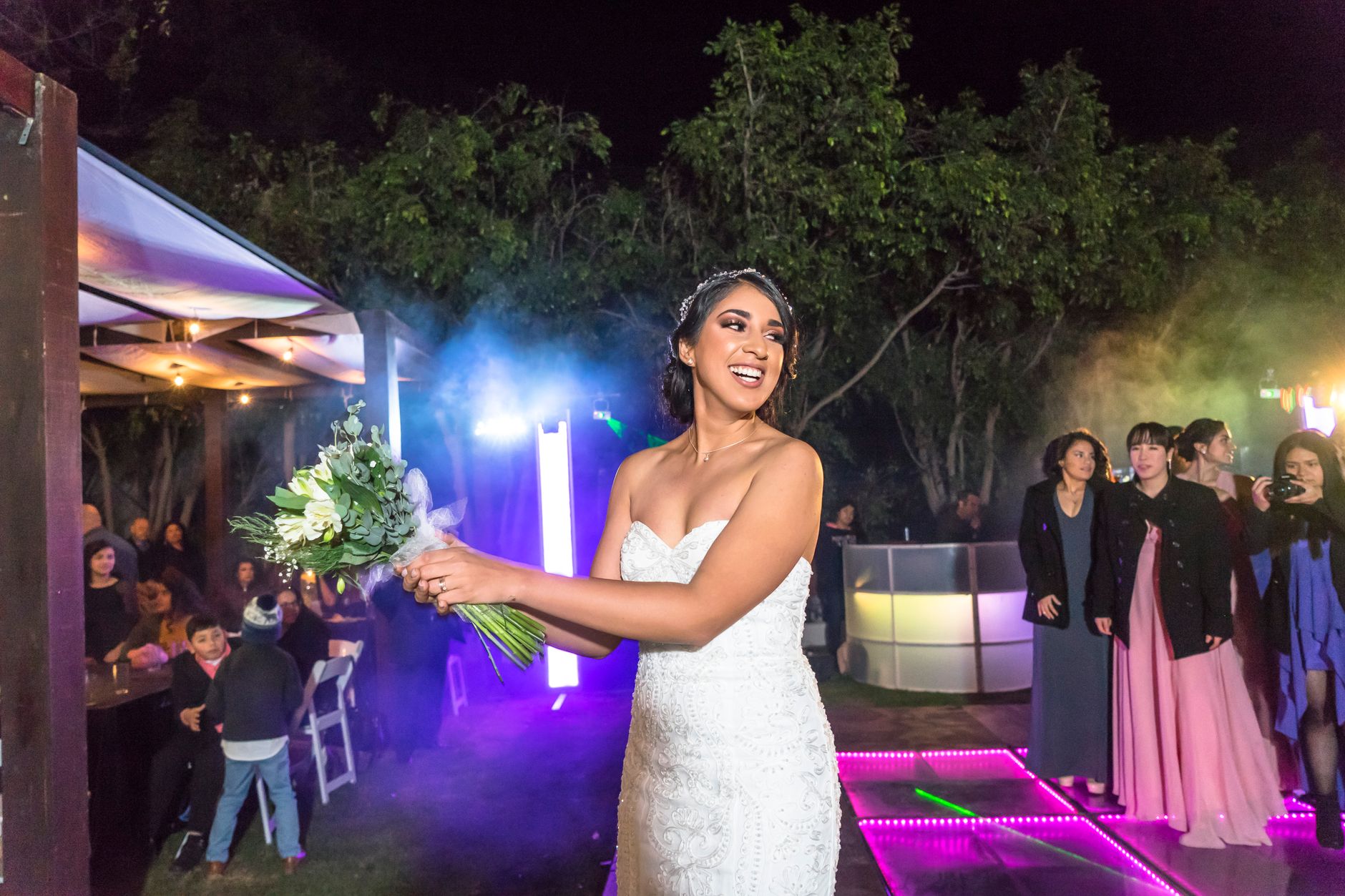 Excited bride in elegant wedding dress prepares to toss bouquet during lively night-time reception.