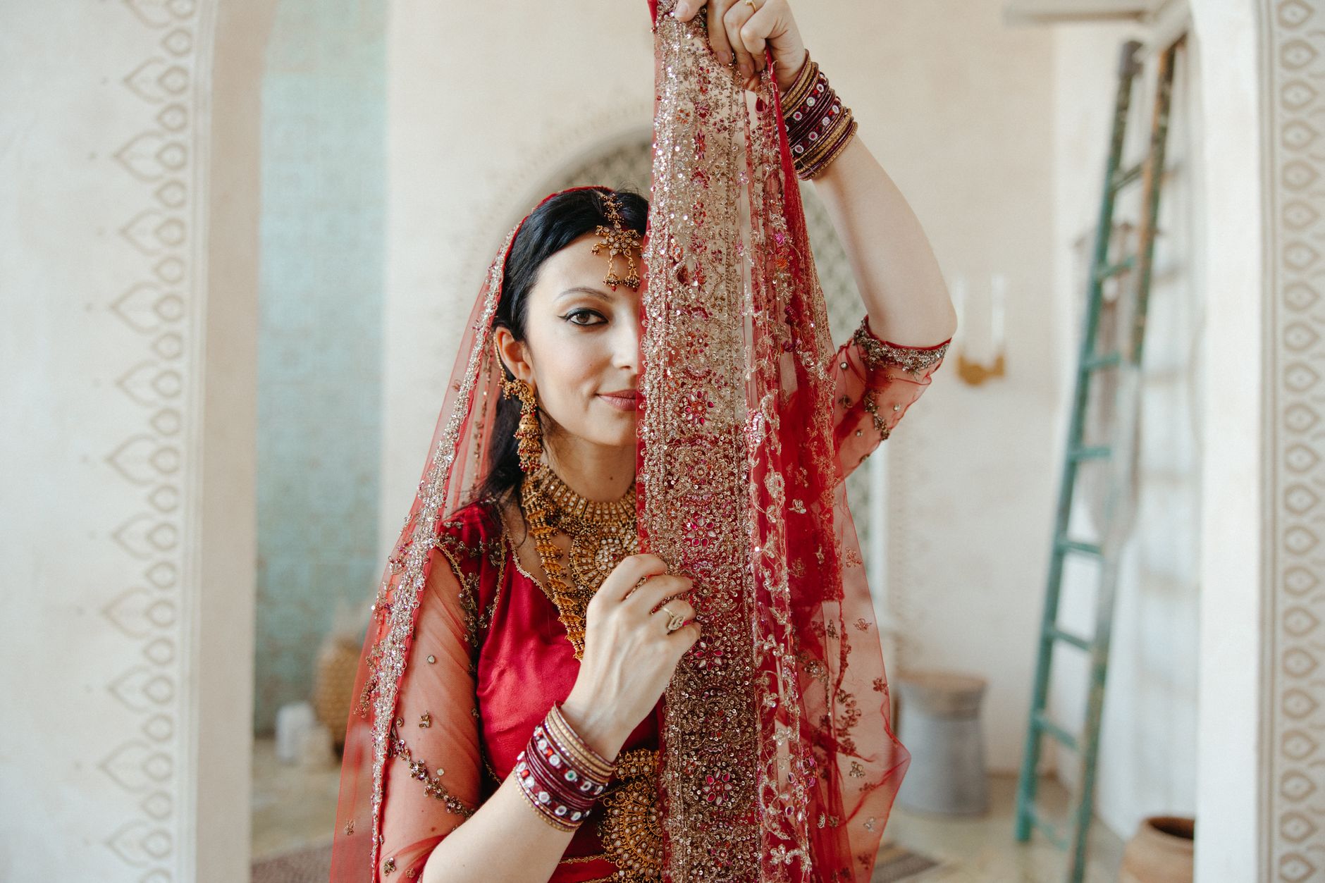 A beautiful Indian bride in traditional red attire posing indoors with intricate jewelry.