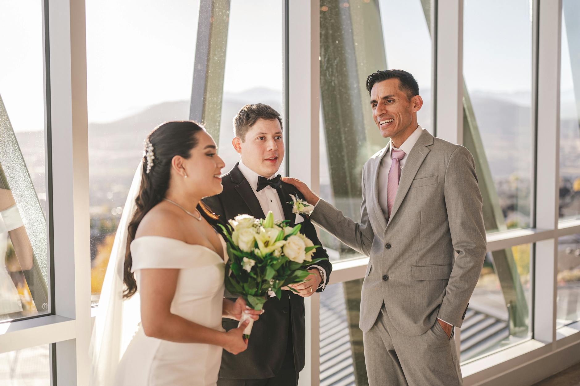 A joyous indoor wedding scene featuring a bride, groom, and best man in elegant attire, set against a bright background.
