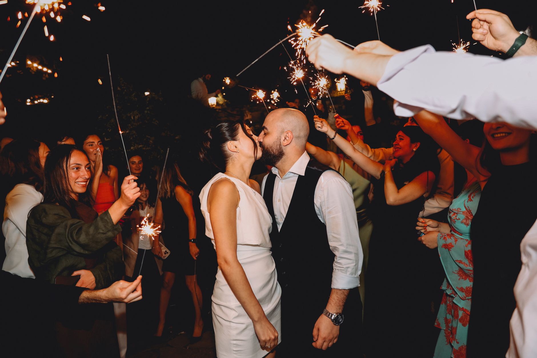 Couple sharing a kiss at night surrounded by sparklers and guests, creating a festive wedding atmosphere.