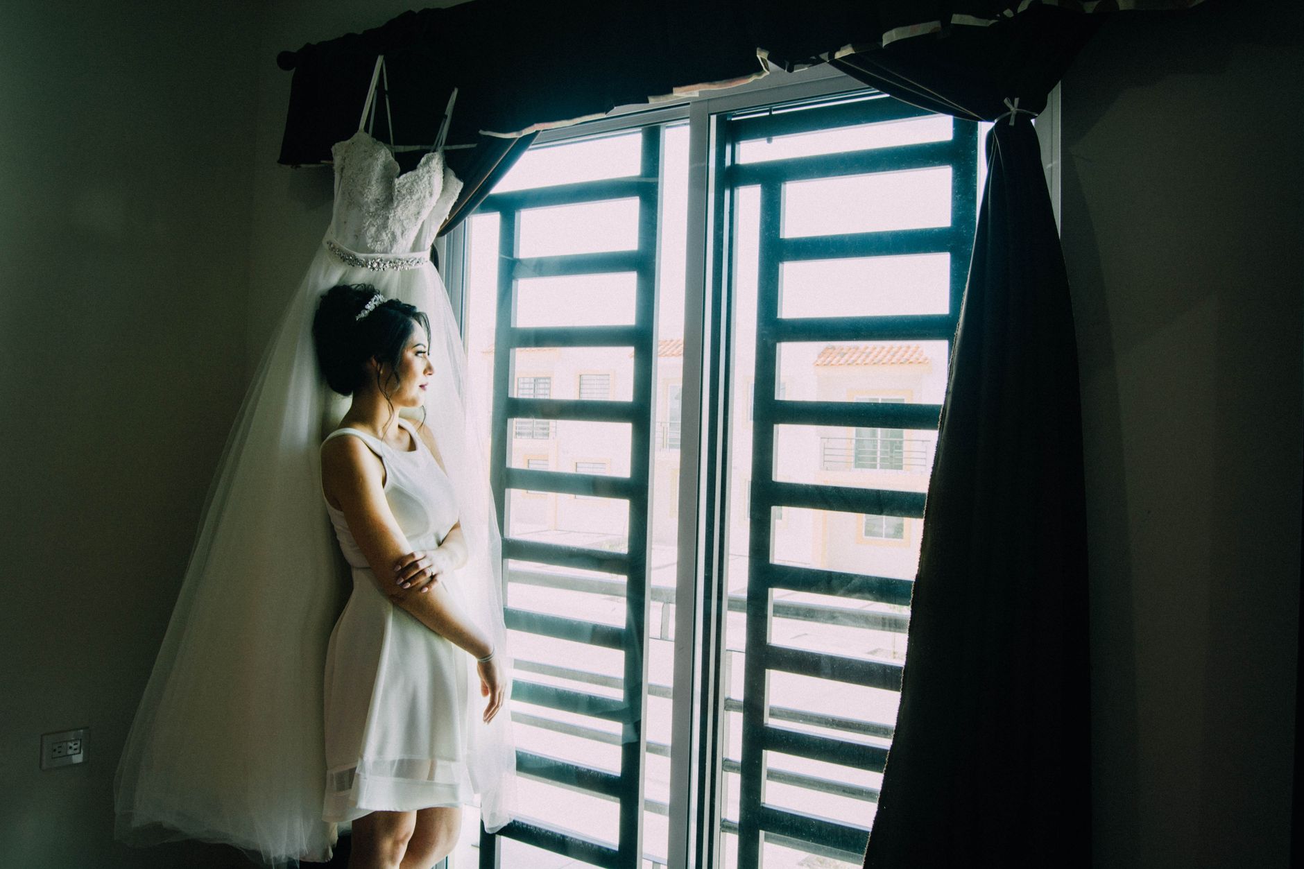 Bride stands by window in white dress, reflecting on her special day. Captures intimate wedding moment.