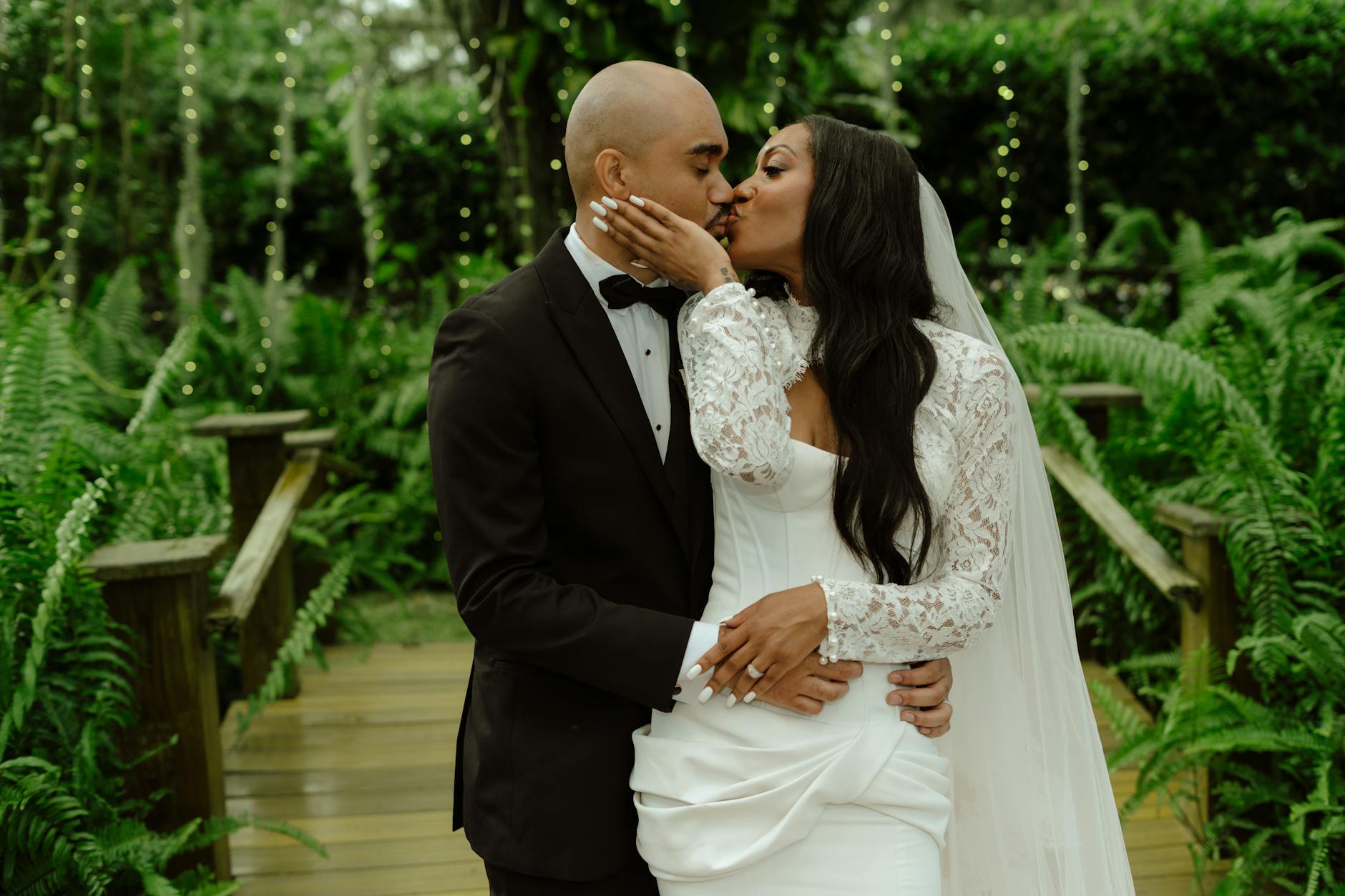 A bride and groom share a romantic kiss on a garden bridge under twinkling lights.