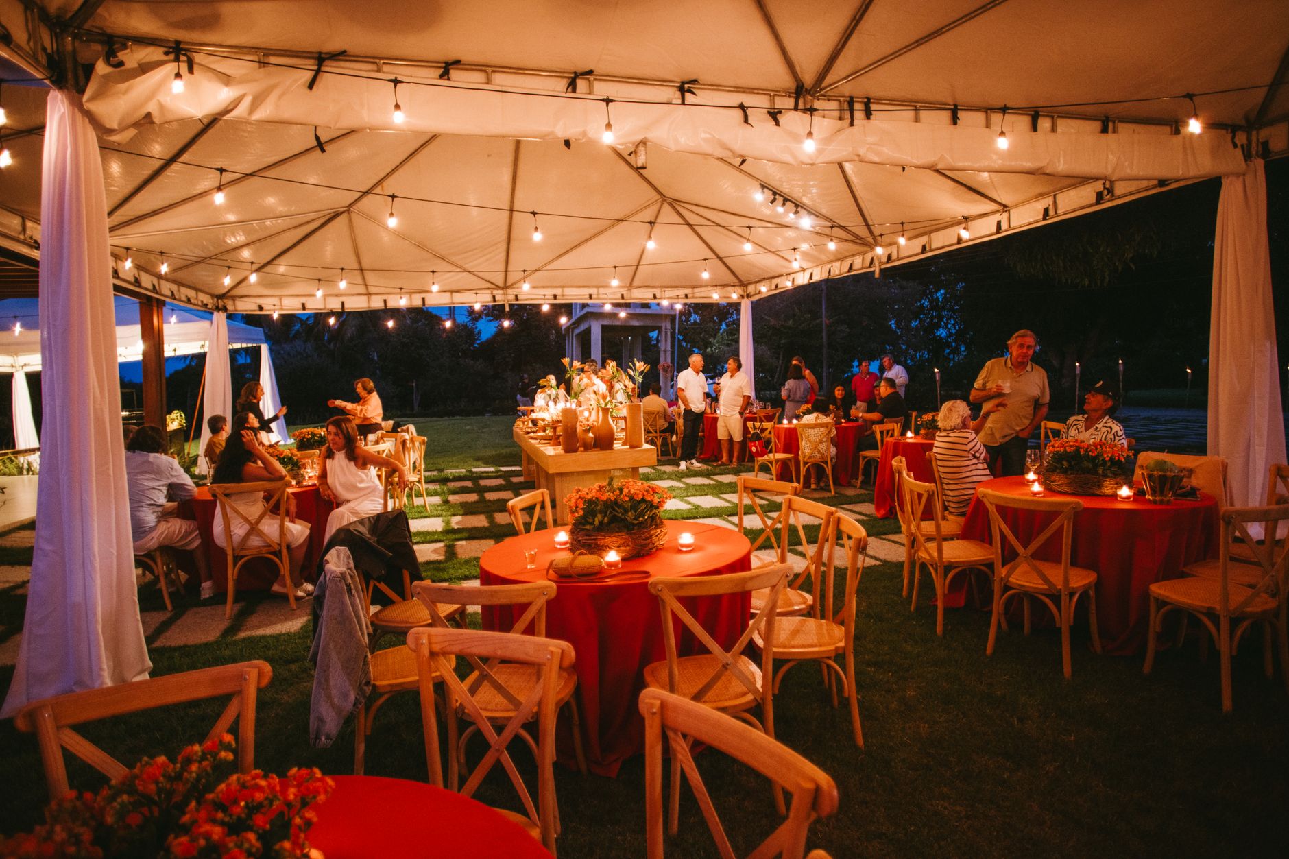 Charming outdoor evening event setup with string lights, red tablecloths, and wooden chairs under a canopy.