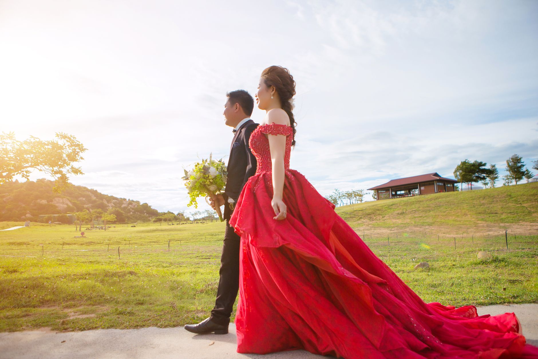 Side view of cheerful ethnic bride in colorful wedding dress strolling on walkway near groom with flower bouquet in rural zone with green hill and house under sky in sunlight