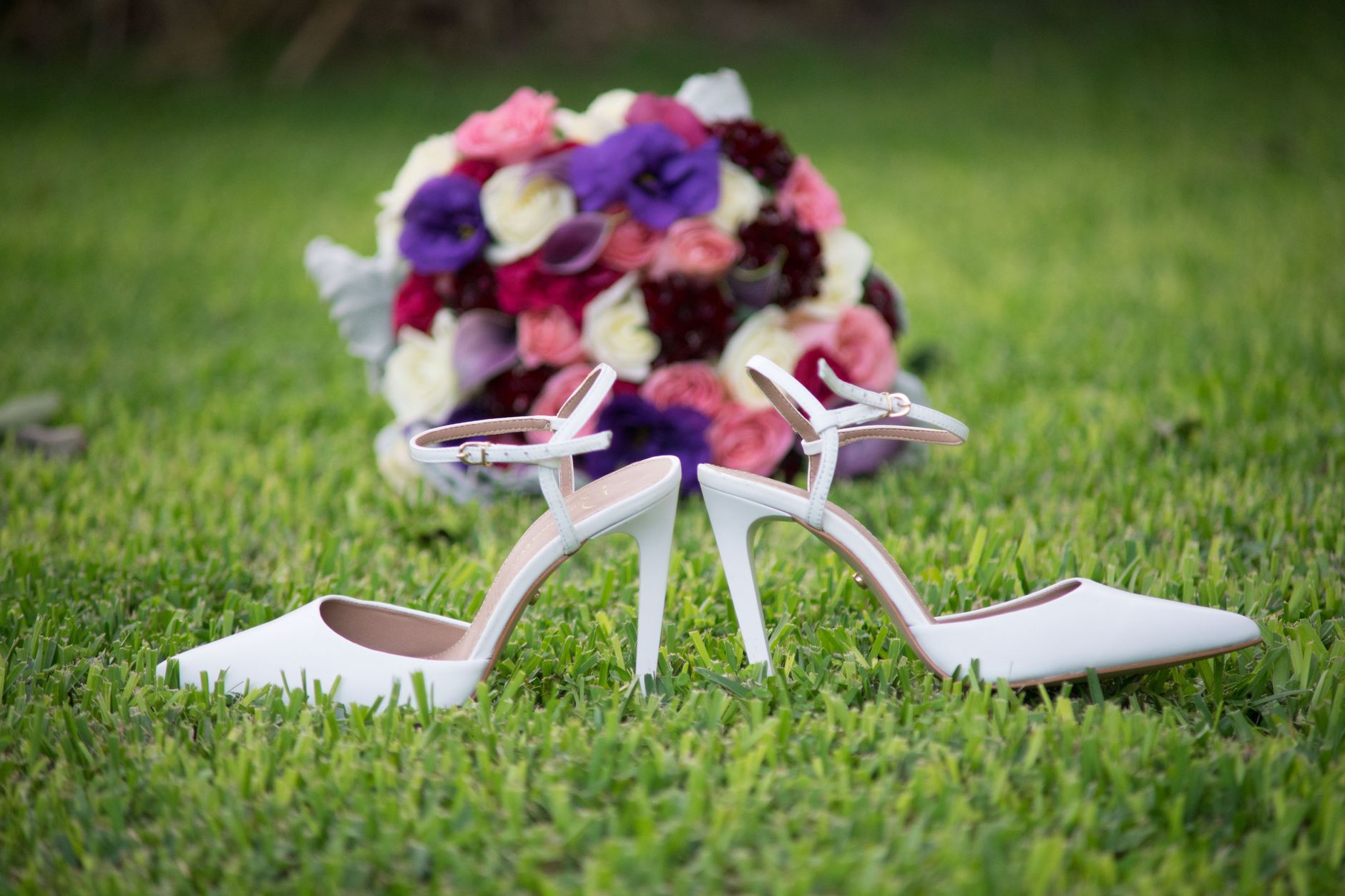 White bridal stilettos and a colorful flower bouquet placed elegantly on a lush green lawn.