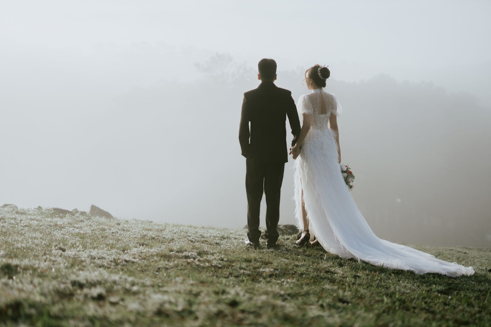 Back view of unrecognizable groom and bride in wedding outfits holding hands and walking on grassy foggy hill in countryside