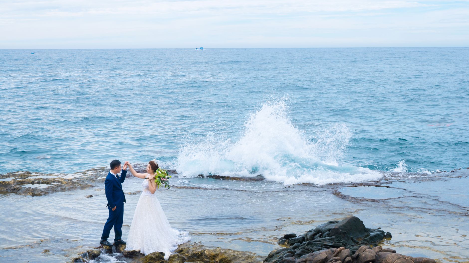 From above side view of anonymous bride in wedding dress with flower bouquet holding hand of groom while standing near sea with splattering water and looking at each other