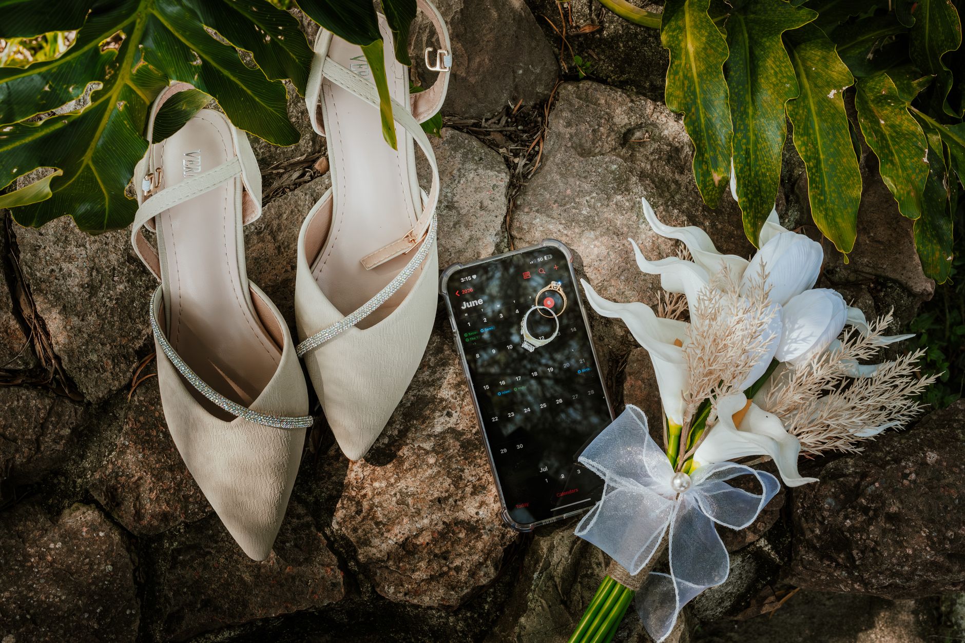 A flat lay of wedding accessories on rocks with shoes, phone, and bouquet.
