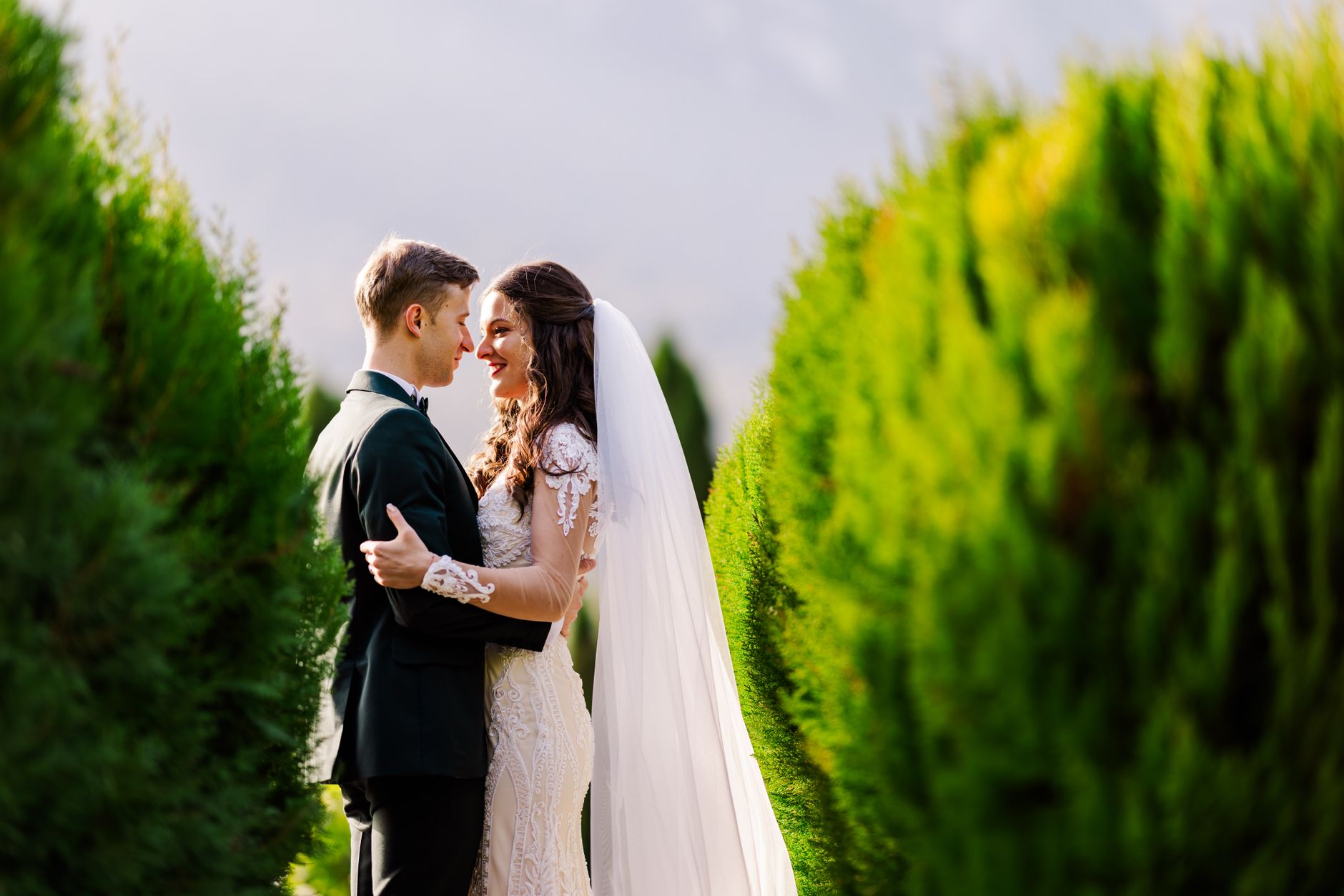 A bridal couple embraces romantically outdoors amidst lush greenery.