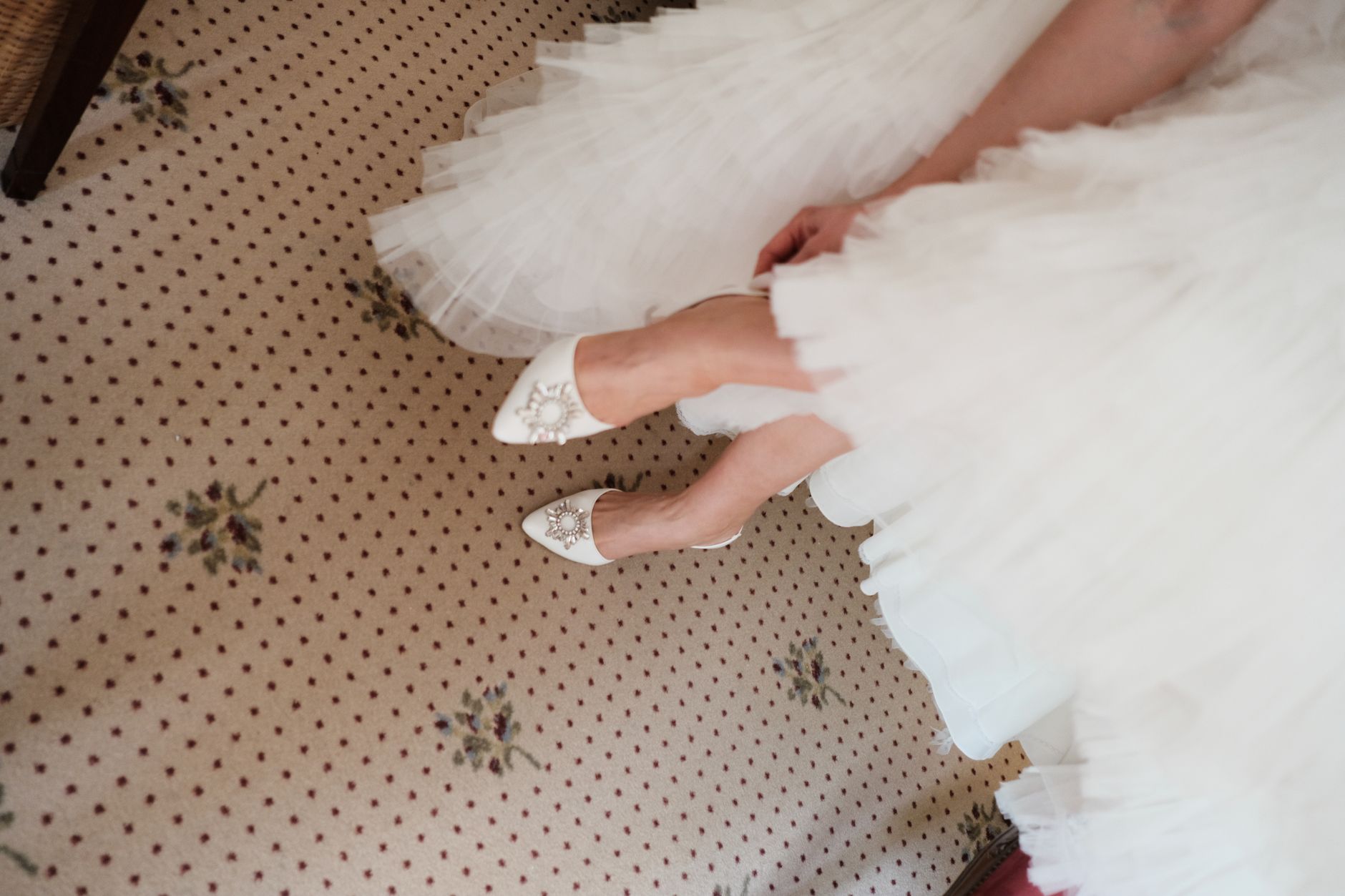 A close-up of a bride in white heels and gown on patterned carpet, ready for a special day.