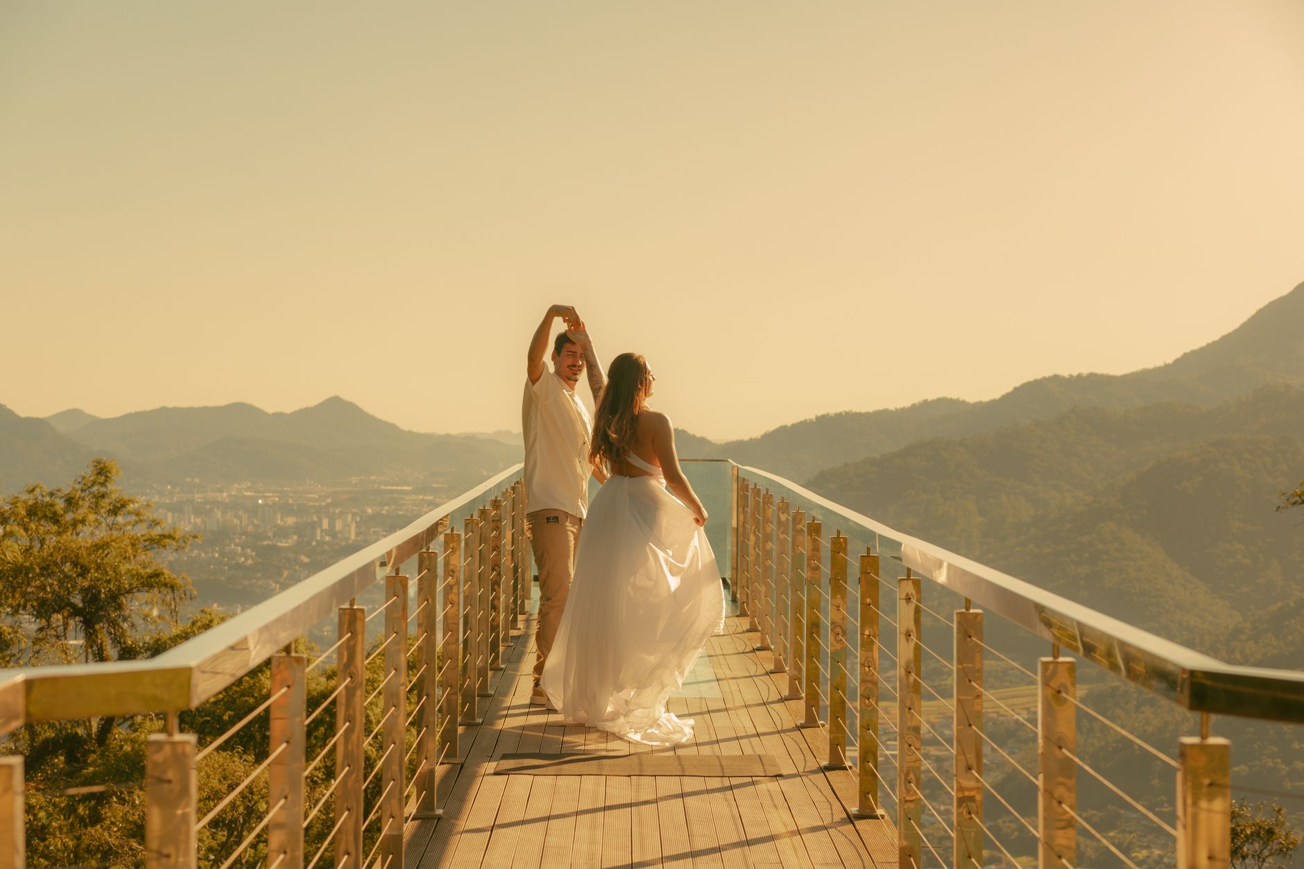 A couple dances joyfully on a scenic wooden deck overlooking mountains and a cityscape under a warm, golden sunset.