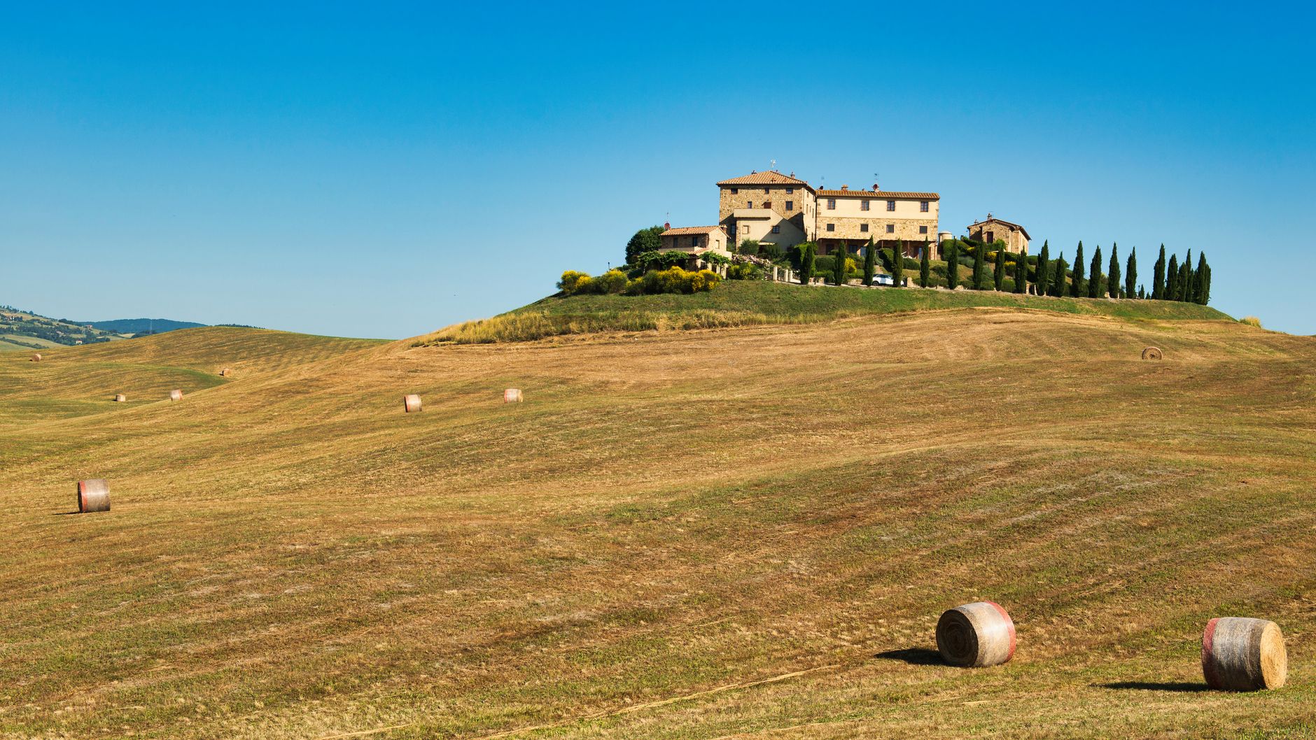 A picturesque Tuscan farmhouse atop rolling hills surrounded by hay bales under a clear summer sky.