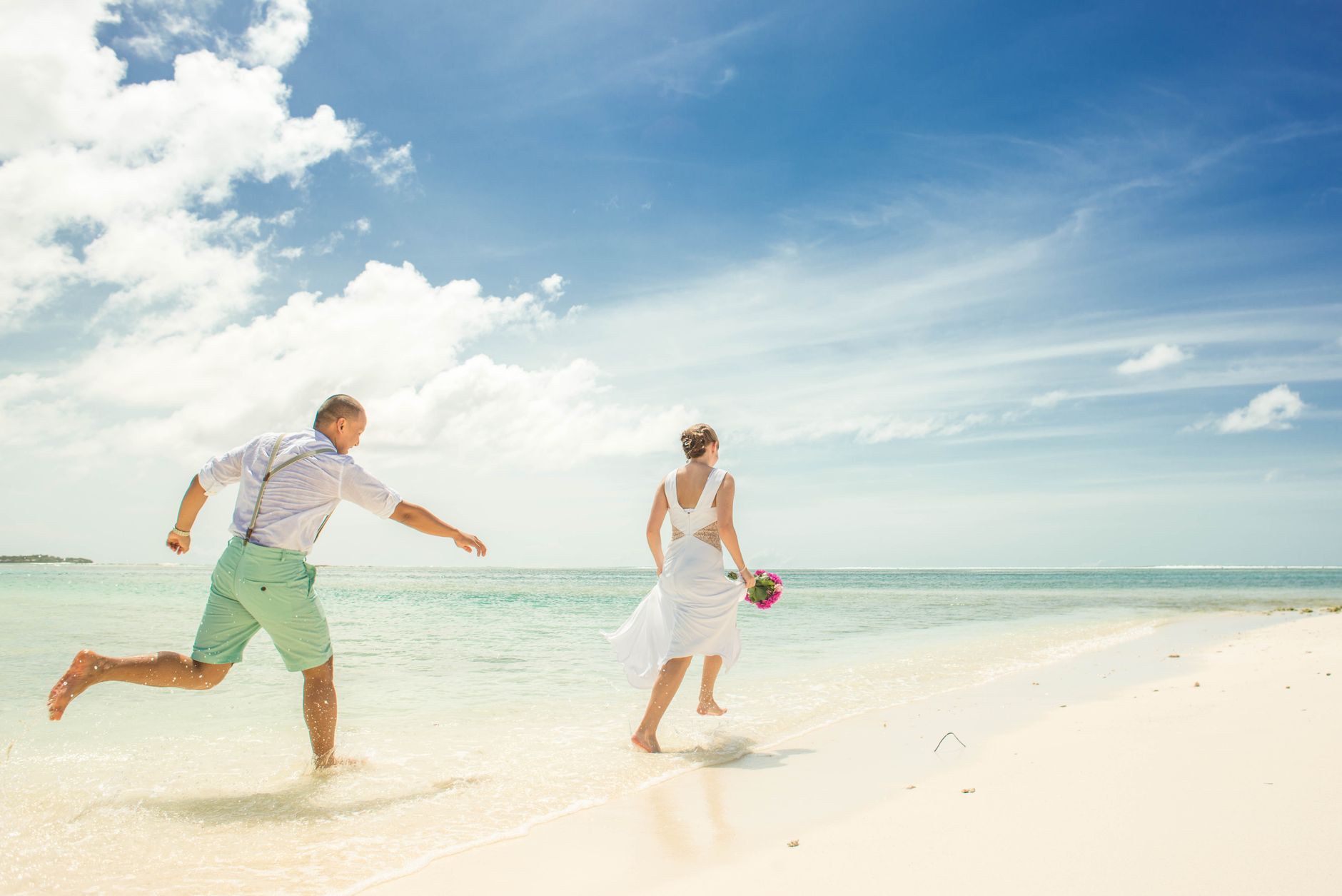 A couple in wedding attire joyfully running along a sunny tropical beach.