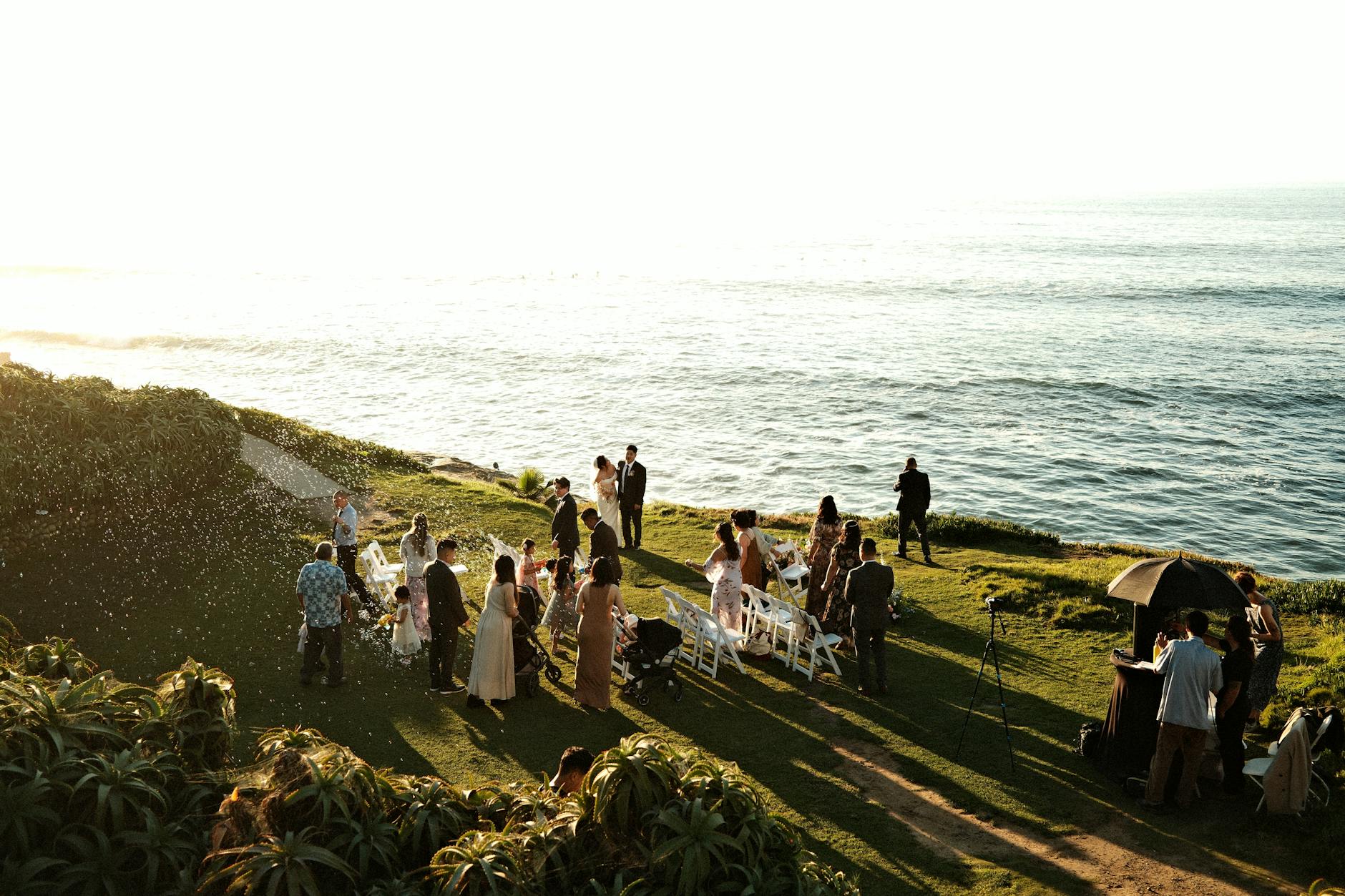 Elegant seaside wedding ceremony with guests gathered on a cliffside during sunset.