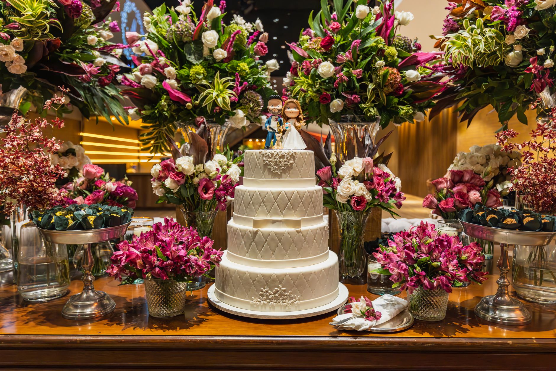 Three-tier white wedding cake surrounded by vibrant flower arrangements indoors.