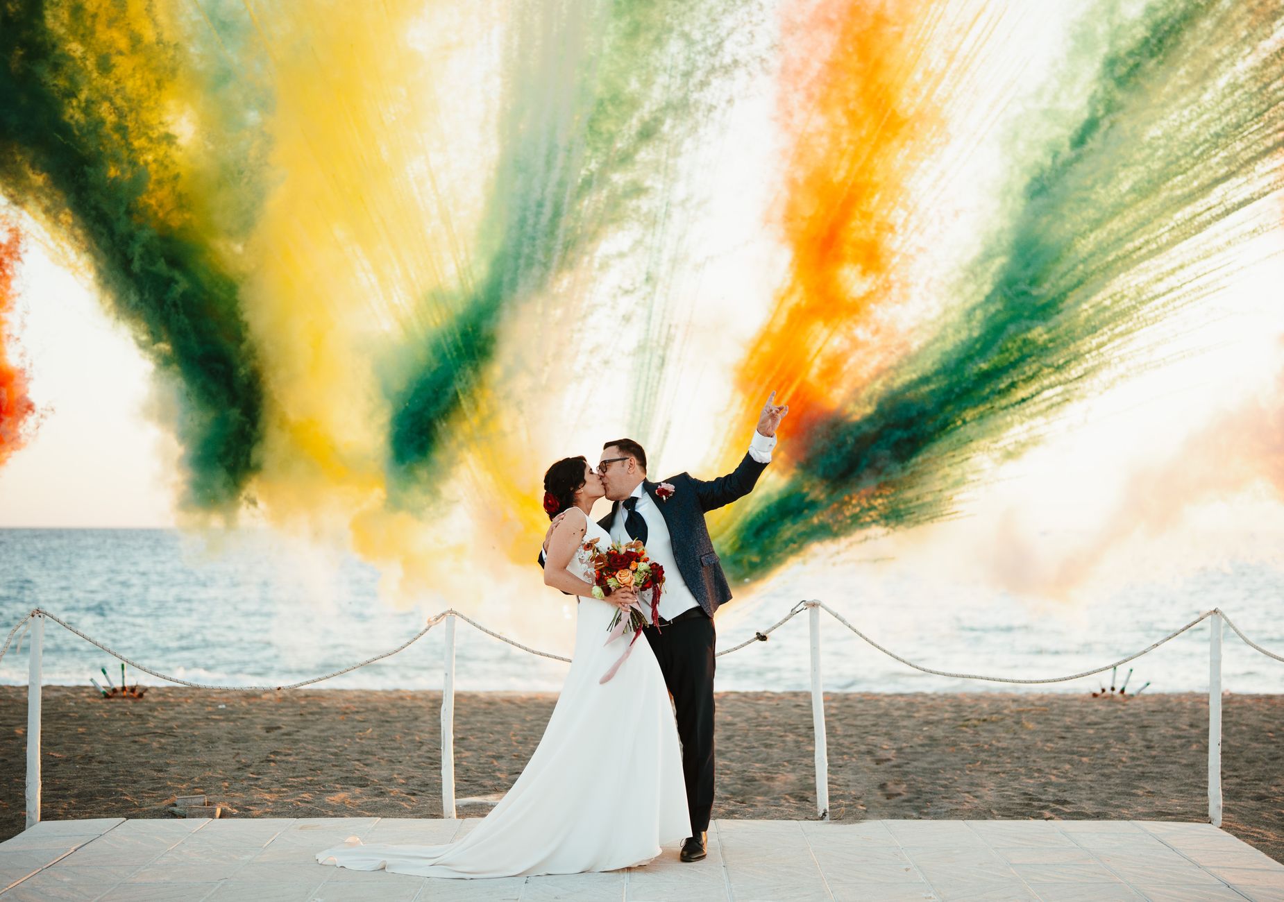 A couple shares a kiss on the beach with vibrant colored smoke creating a stunning backdrop.